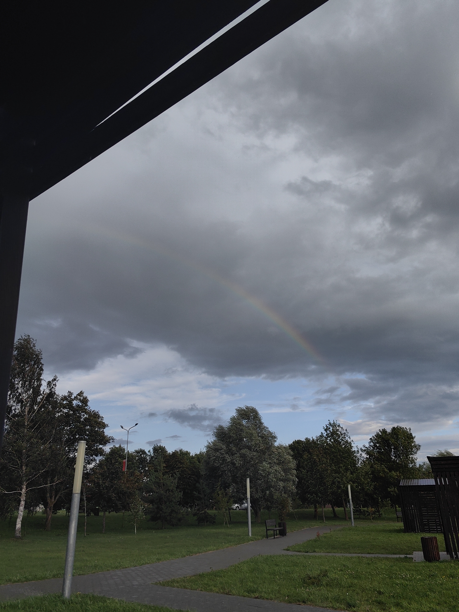 The image depicts a park scene in Myadzyel, Belarus, under a cloudy sky. A faint rainbow is visible in the upper portion of the frame, arcing through the dense, grey clouds. In the foreground, a dark, angular structure, possibly a part of a building's overhang or canopy, partially obscures the view of the sky. A paved pathway winds through a grassy area dotted with trees. A lamppost stands to the left of the path, and a bench with a trash bin is situated further along. In the background, more trees and some dark wooden structures, potentially gazebos or sheds, are visible. The overall atmosphere is subdued due to the overcast conditions.