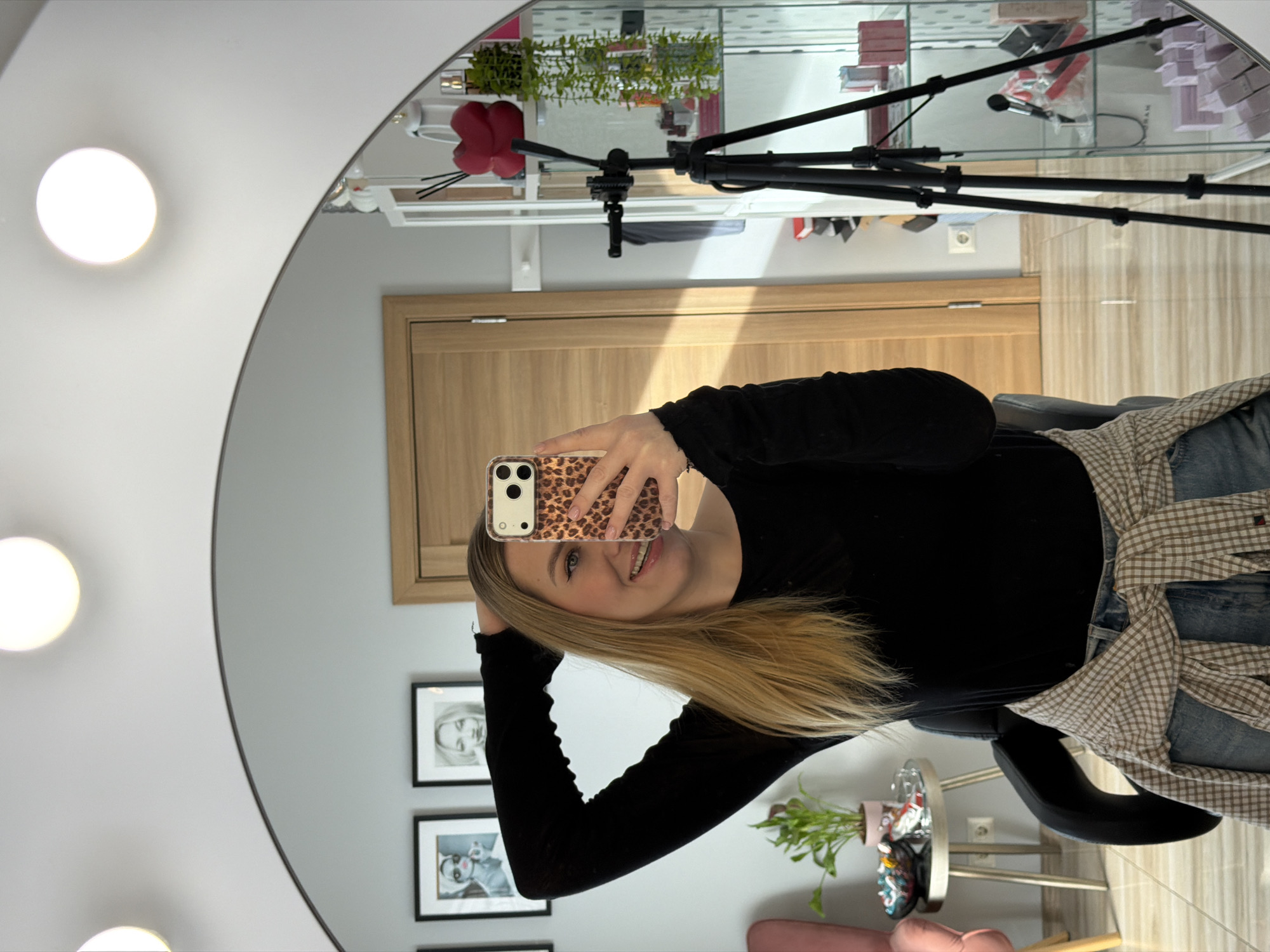 A medium shot shows a blonde woman taking a mirror selfie with an iPhone in what appears to be Riga, Latvia, inside a room adorned with a lighted mirror. The woman is wearing a black long-sleeve shirt, jeans, and a patterned shirt around her waist. Her phone has a leopard print case. She is smiling and looking directly at the camera. In the background, there's a door, framed pictures, a lighting setup, plants, and shelves displaying various items. A make-up table with a round mirror and bulbs is noticeable. The mirror reflects items such as a hair styling product, decorative elements, and a red heart prop. The environment suggests a professional setting, possibly a beauty salon or a makeup studio.