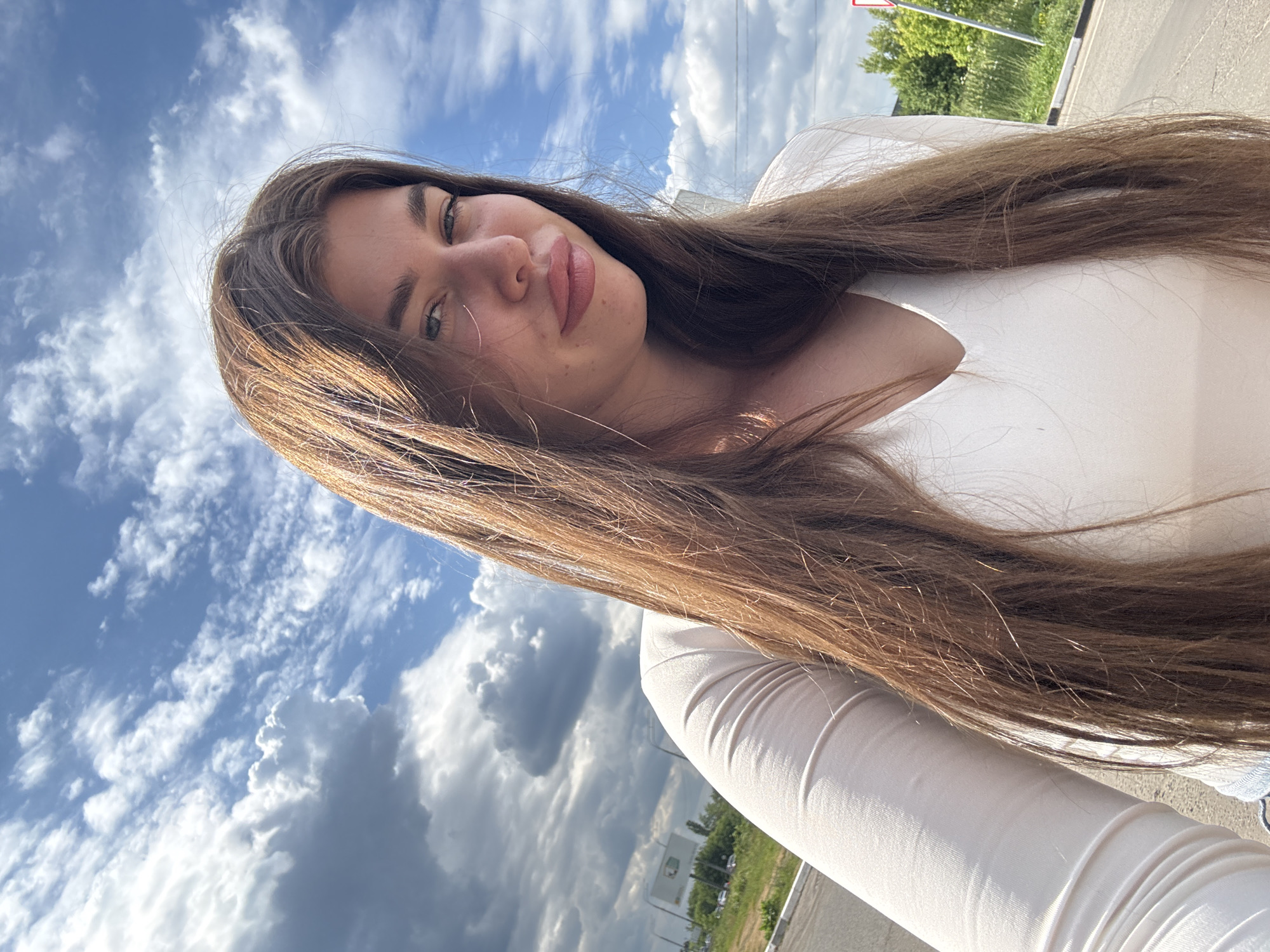 A young woman with long, brown hair takes a selfie outdoors. She is wearing a white, long-sleeved top. Her hair is flowing around her face and shoulders, obscuring parts of her body. The background is a cloudy blue sky. Sunlight illuminates her face, highlighting her skin and makeup, which includes lipstick. She appears to be smiling or smirking at the camera.
In the background, beyond the sky, there are hints of a roadside setting. Green trees and a pole with a sign are visible on the right side of the image, suggesting an outdoor environment near a road. The lighting indicates it is daytime, likely a sunny afternoon given the brightness and the presence of clouds. The clouds themselves are a mix of white and gray, with some appearing fluffy and others more diffused, suggesting a dynamic weather pattern.
No visible text is present in the image. The overall mood is casual and carefree, consistent with a selfie taken on a pleasant day. The orientation of the photo is portrait, but the image itself is rotated, with the subject's head appearing at the top left of the frame.