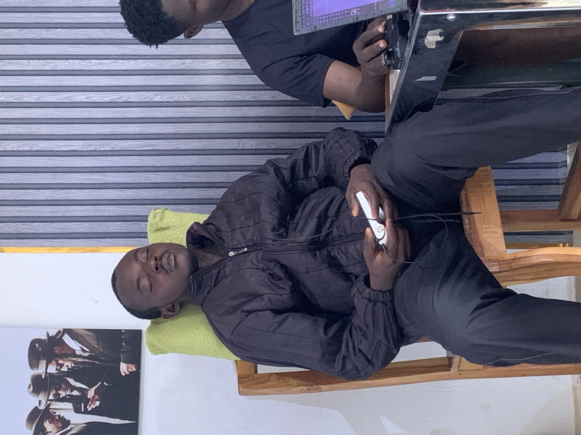 A man is reclining in a wooden chair with a green pillow under his head, appearing to be sleeping. He is wearing a dark jacket and pants and holding a white earbud-style headphone in his hand. Another man is seated in front of him, operating a piece of electronic equipment with a screen. The setting is indoors, with a picture of men in hats on the wall and a striped background behind the men. I cannot determine the location in the city of Jos, Nigeria from this image.