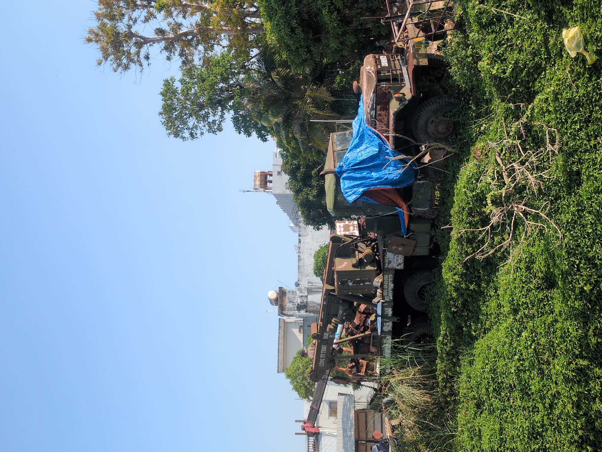 The image shows a roadside scene in Tuy Hòa, Vietnam. An overgrown, derelict military-style truck covered in a blue tarp sits amidst dense green vegetation. In the background, there is a building with a satellite dish and tower, typical of the urban landscape of Tuy Hòa. A construction crane is visible on the lower left, indicating building activity in the area. The sky is clear blue, suggesting fair weather conditions.
