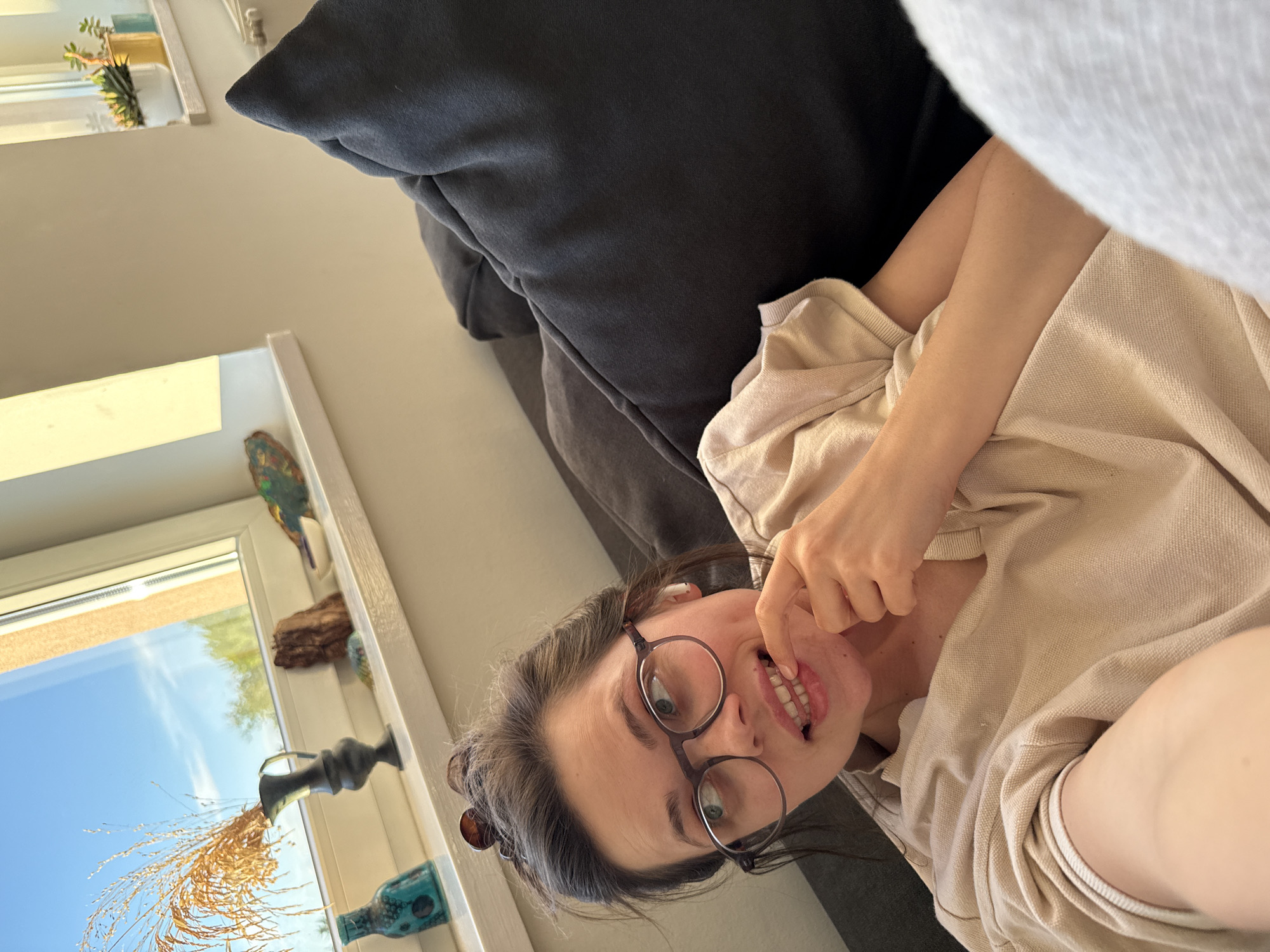 A young woman with dark hair and round-framed glasses is captured in a close-up, angled shot, reclining on a dark grey couch indoors. She wears a light-colored (beige/cream) collared shirt or hoodie and has an earbud in her left ear. Her expression is playful, with a finger touching her upper lip, revealing her teeth as she looks directly at the camera.
The background shows a white-framed window through which a clear blue sky with sparse clouds is visible, suggesting a sunny daytime. On the windowsill, several decorative items are arranged: a small green plant in a white pot, a piece of wood, a dark metallic vase holding dried golden grasses, and a teal/green decorative bottle. The natural light illuminates the scene, highlighting a relaxed and casual atmosphere. No text is visible in the image. The location context provided is Budapest, Hungary.
