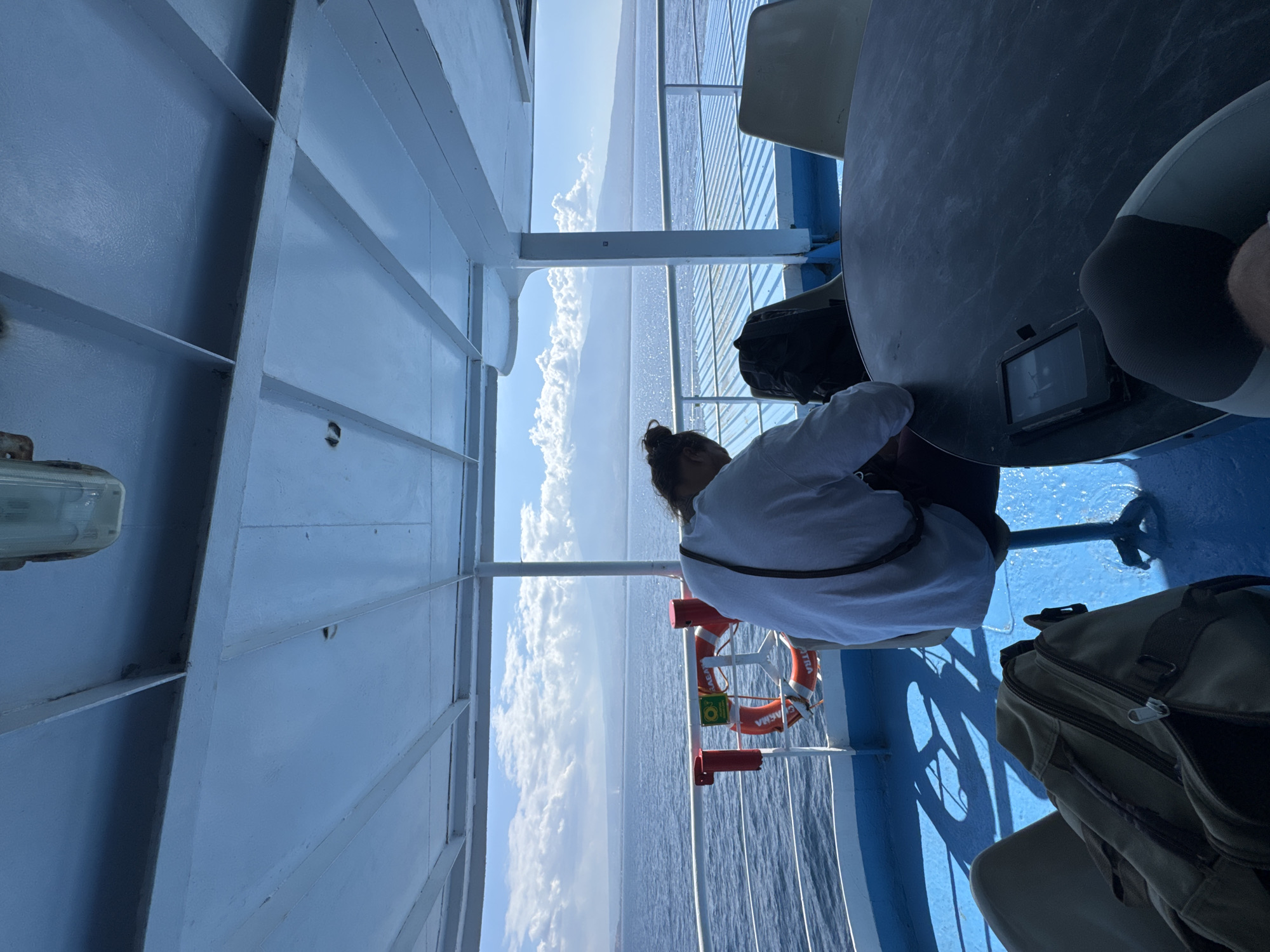 The image captures a seated person on a ferry with a view of the ocean and a distant island. The person, viewed from behind, is wearing a white long-sleeved shirt and has their hair in a bun. They are sitting at a dark circular table with a black backpack next to them. A tablet is also visible on the table.
In the foreground, a large duffel bag sits on the blue deck. In the middle ground, a life preserver with orange and white coloring is attached to the railing. Beyond the railing, the vast blue ocean stretches towards a hazy shoreline with a mountainous landscape under a sky filled with white clouds. The ferry's white interior structure, including beams and a light fixture, is visible overhead. The blue deck, white railing, and the ocean are the dominant colors in the scene. The overall impression is one of travel and contemplation during a ferry journey.
The location is identified as being on a ferry in Unknown, Indonesia.
