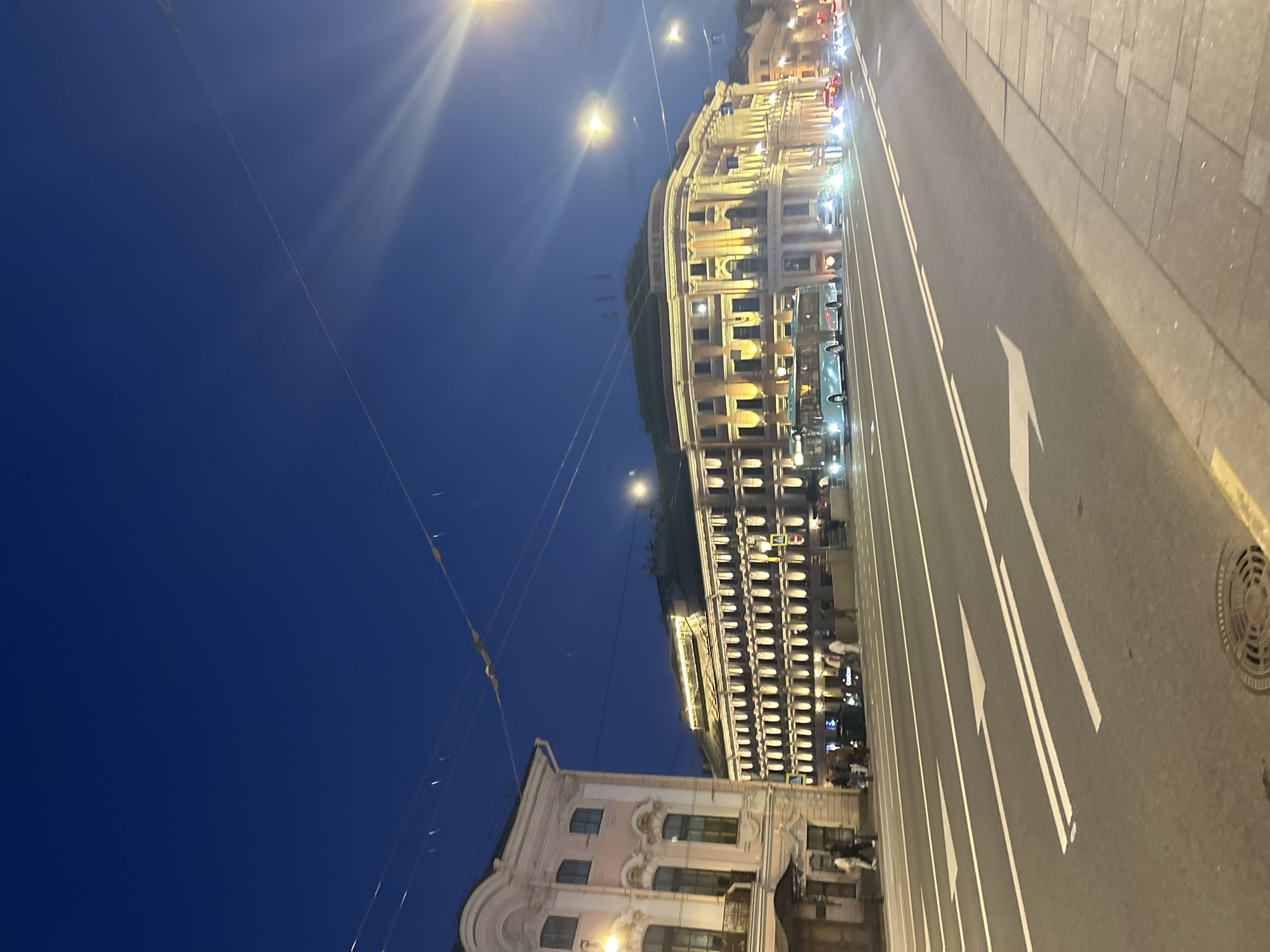 An outdoor, eye-level, medium shot captures a wide street in an urban setting at dusk. The sky is a deep, dark blue, with a few faint lights visible. Overhead wires crisscross the scene.
On the left side of the street, a pale pink building with ornate details and balconies stands tall. A few people are visible walking on the sidewalk in front of it.
Dominating the center and right of the frame is a grand, multi-story building illuminated with warm yellow lights, highlighting its architectural features. Rows of windows are clearly visible, and lights glow from within. In front of this building, several cars and a light blue bus are on the road. The bus appears to be in motion.
The street itself is paved and marked with white lane lines and directional arrows. A sidewalk lines the right side of the road, separated by a curb. A circular manhole cover is visible on the road in the lower right corner of the image. The overall impression is of a bustling city street at the beginning of the night, with illuminated architecture and active traffic.
