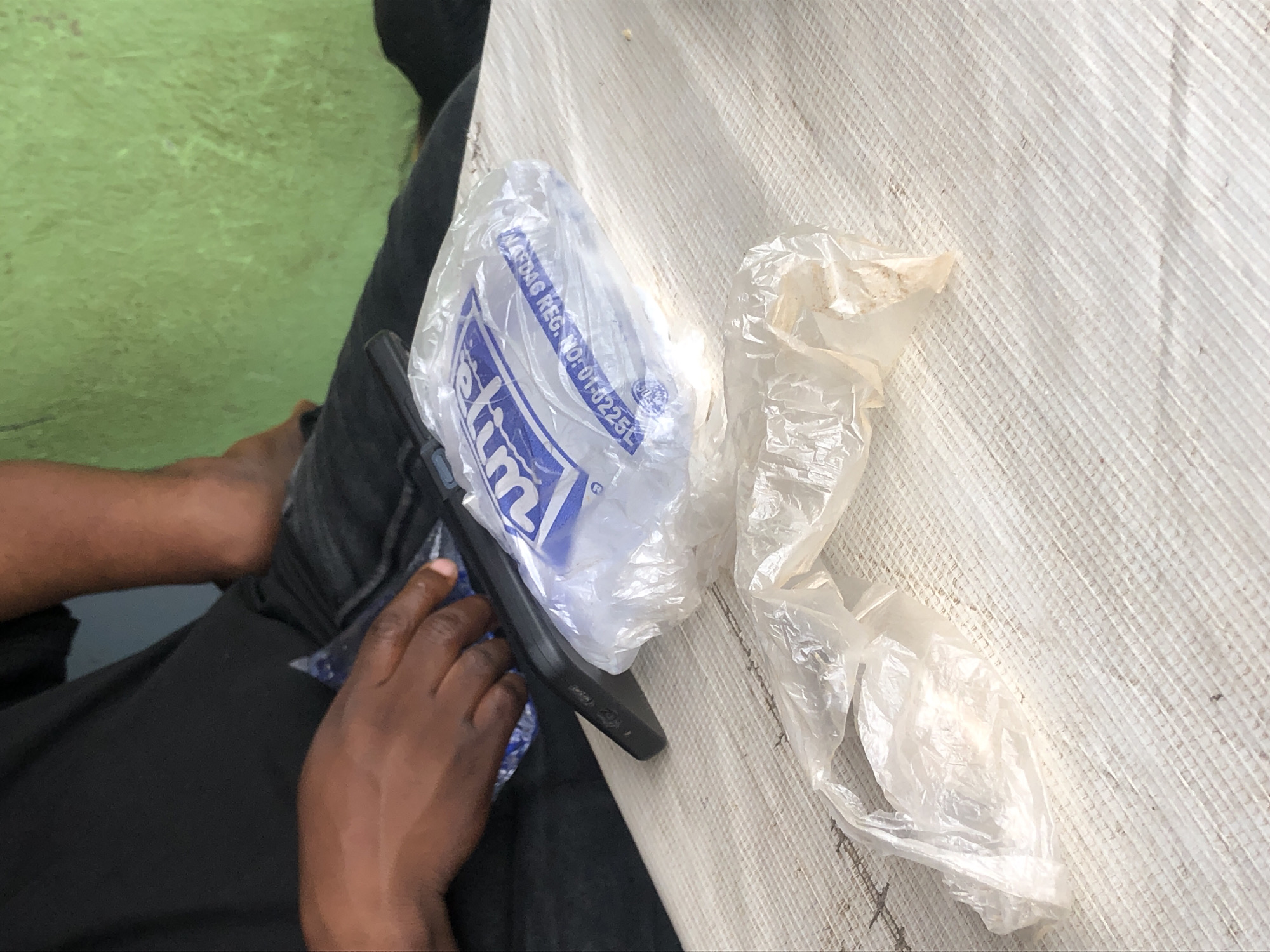 A close-up shot captures a person's hands holding a phone and a plastic bag on a textured white surface. The person is wearing a black t-shirt and dark pants. In the background, a green wall is visible. The plastic bag contains some white powdery substance. The overall scene appears to be in an outdoor setting in Kabong, Nigeria.