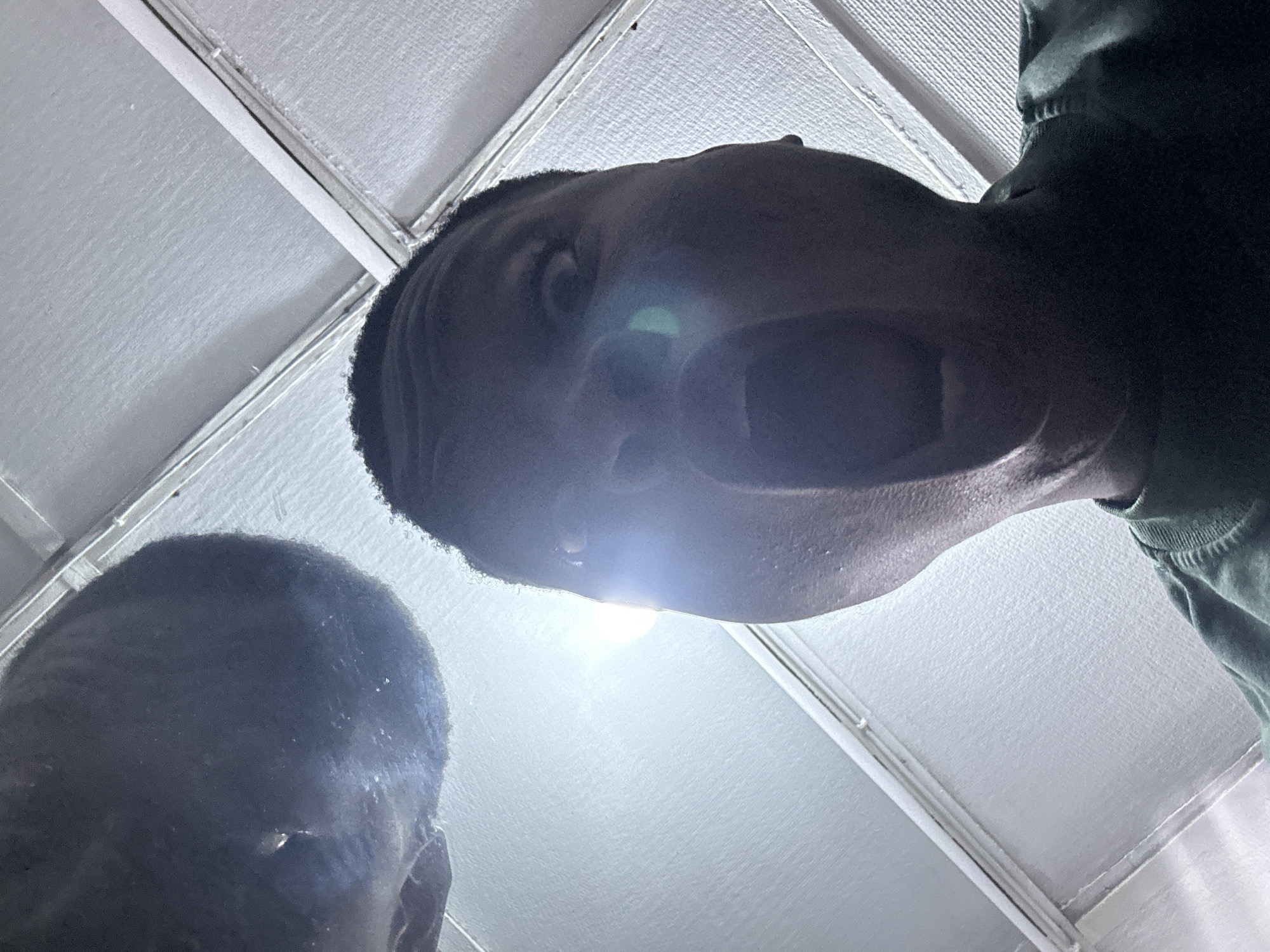 The image captures two individuals, likely Nigerian, viewed from a low-angle perspective looking upwards. The prominent subject, positioned in the upper right, has dark skin and short dark hair. Their eyes are wide open, and their mouth is agape in a broad expression, suggesting a shout, laugh, or intense reaction. A strong, artificial light source directly beneath their face creates significant lens flare and dramatic underlighting. On the lower left, the top of a second individual's head and a small portion of their face are visible, also with dark skin and short hair, looking downwards. The background consists of a light-colored, textured ceiling, possibly acoustic tiles, with visible linear elements that could be support beams or panel divisions. The scene takes place indoors, with the time of day being indeterminate due to the artificial lighting. The overall impression is one of intense emotion or a loud vocalization from the main subject, looking down towards the camera's position. There is no visible text in the image. The individuals are likely situated in an indoor setting in Ikorodu, Nigeria.