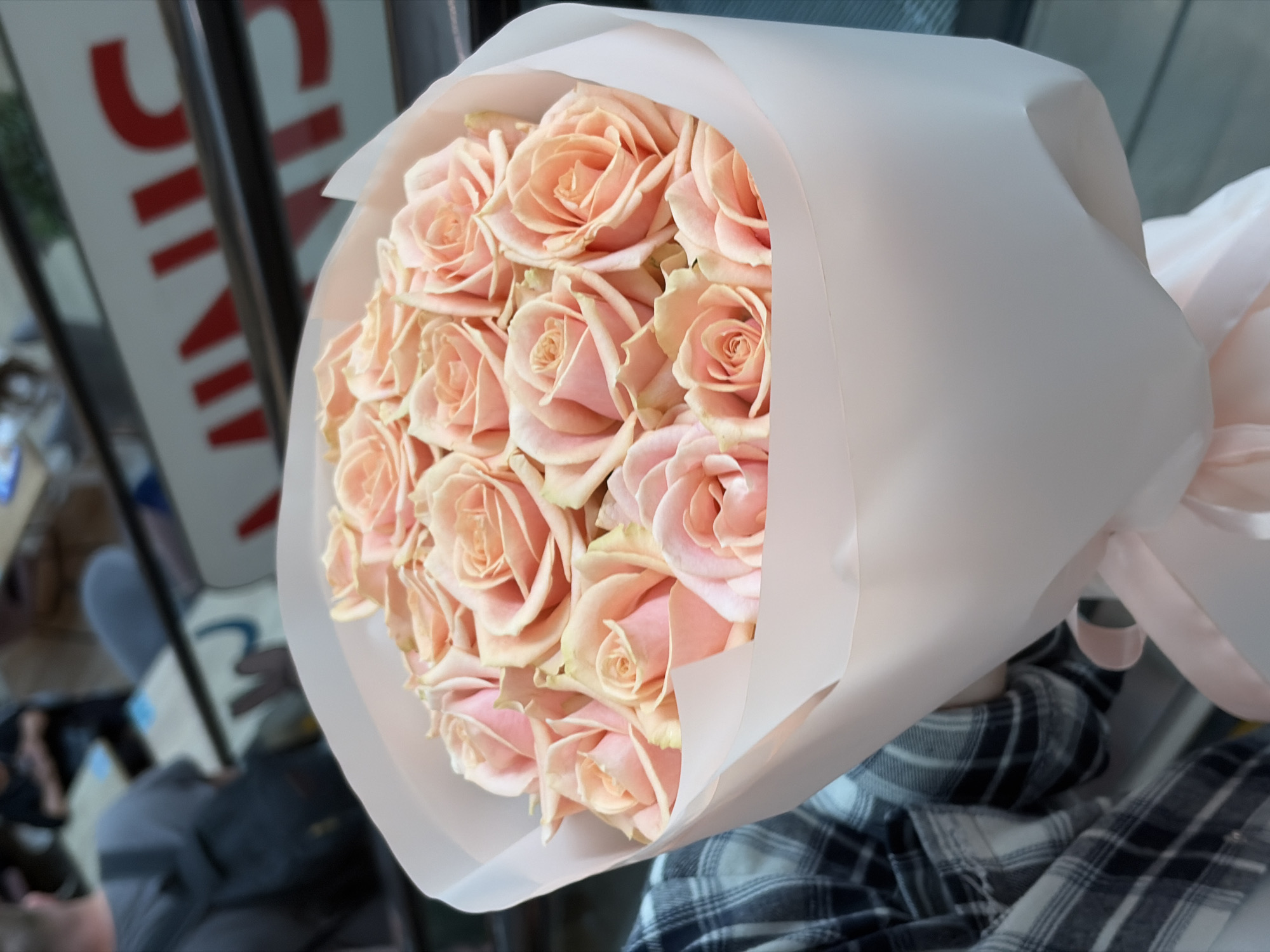 A bouquet of approximately 15 light peach-colored roses is wrapped in white paper and tied with a light peach-colored ribbon. The bouquet is held in front of a blurred background that suggests a city street. A person's arm, clad in a black and white checkered shirt, is visible holding the bouquet. The blurred background reveals a sign with red letters, and hints of other urban elements, possibly indicating a location in Minsk, Belarus.
