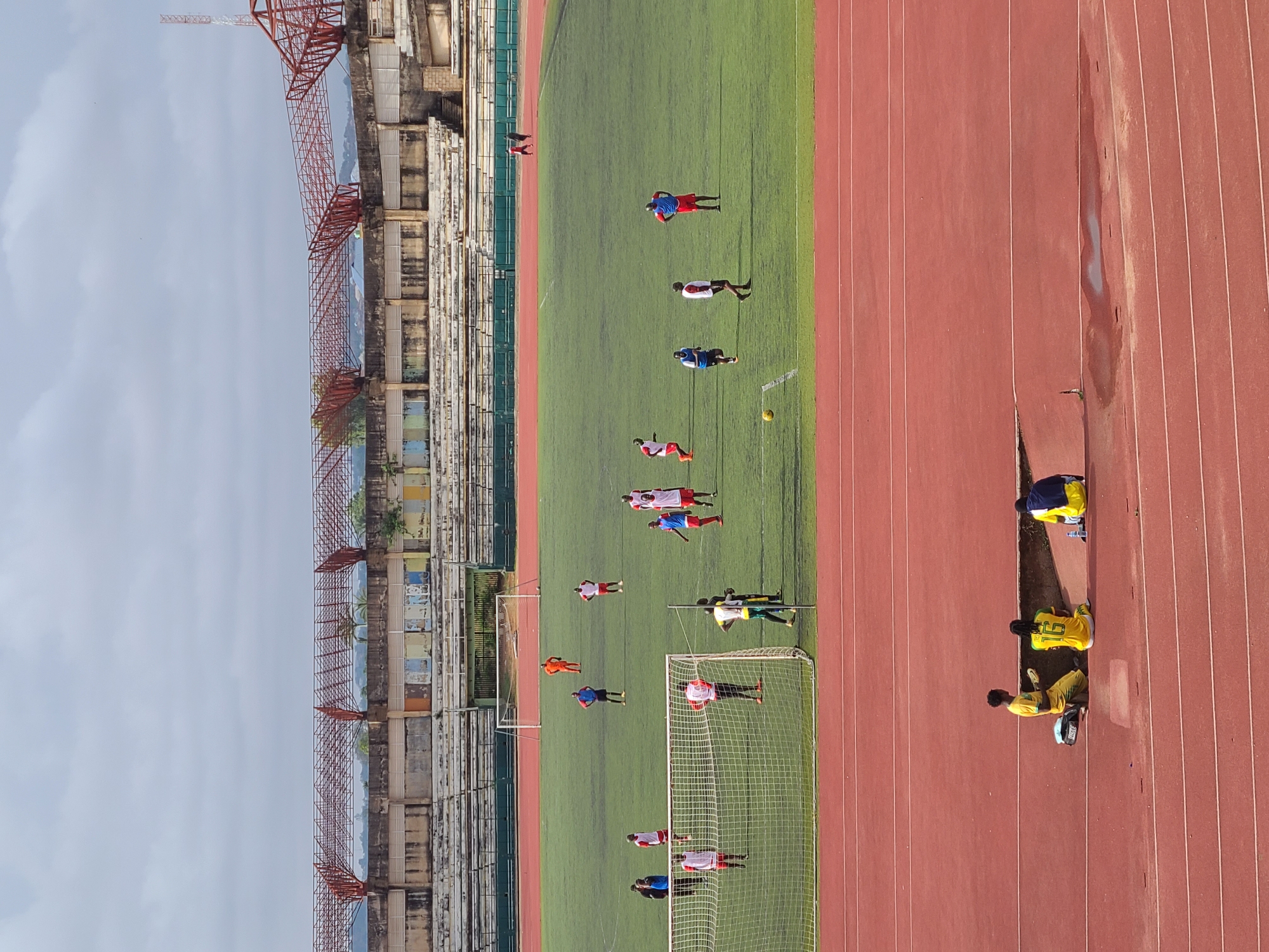 A stadium in Unknown, Unknown, with a track field and green playing field, where players are practicing. The stadium is old and rundown. There is a cloudy sky overhead. The field appears to be damp.