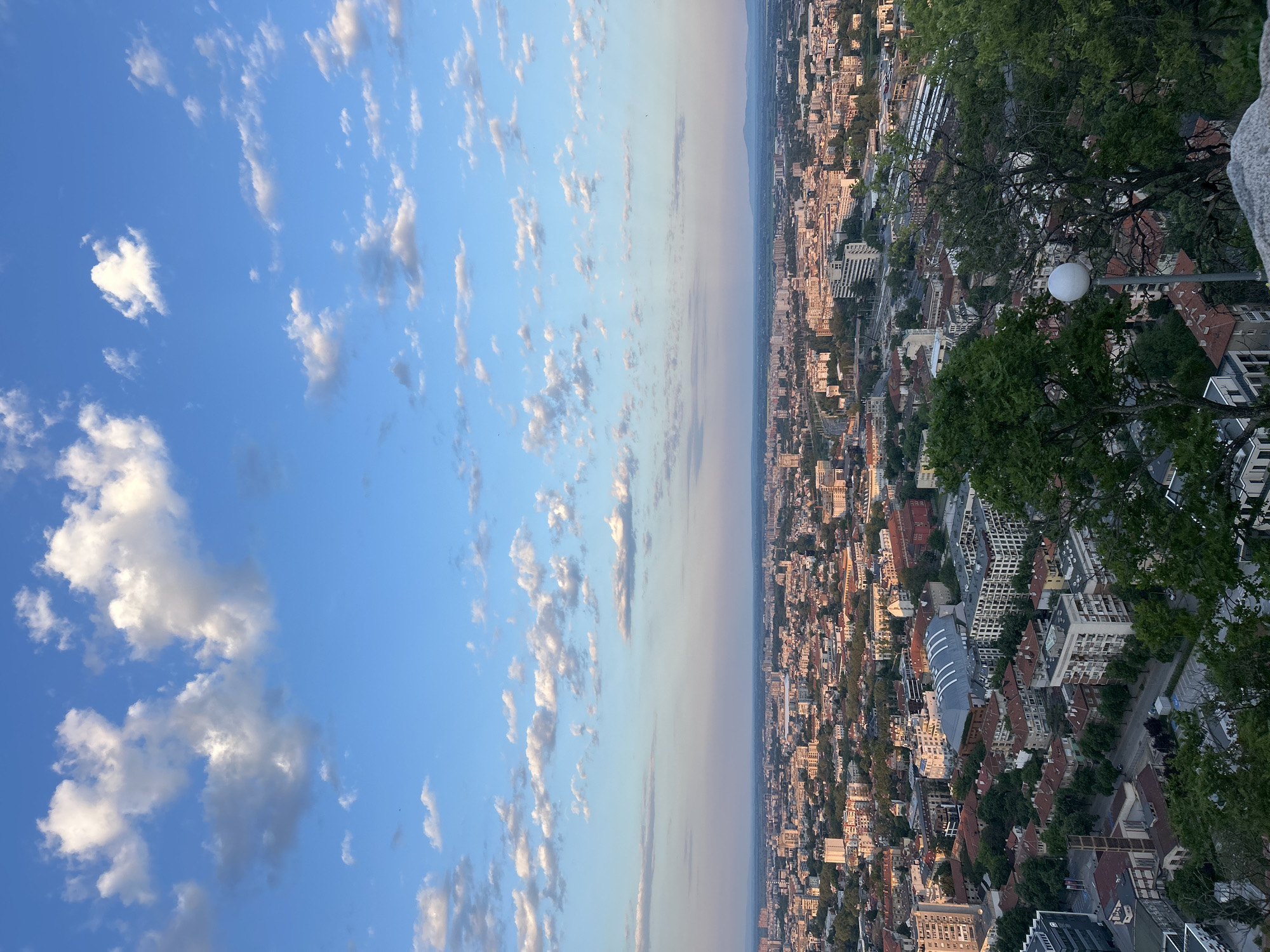 The image presents a high-angle, eye-level view of Plovdiv, Bulgaria, from Nebet Tepe, a historical hill landmark, under a partly cloudy sky. The city sprawls below, densely packed with buildings varying in height and architectural style. The left side of the image is dominated by a blue sky dotted with scattered cumulus clouds. The horizon line, slightly blurred, separates the sky from the cityscape. The right edge includes trees and a streetlight, suggesting an observation point. A street is visible among the buildings. There are no visible people or interactions within the frame.