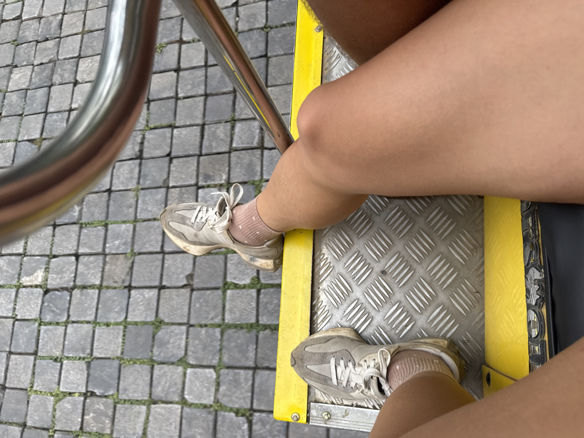 The image shows a close-up perspective of two pairs of legs, clad in white sneakers and light-colored socks, positioned on a metal platform with a yellow edge. The platform appears to be part of a ride or vehicle, indicated by the presence of a metallic railing in the upper right portion of the frame. The ground beneath is paved with cobblestones, interspersed with small patches of grass. One person's legs are more prominently featured, while the other pair is partially visible. The setting does not provide any specific visual cues that would allow for the identification of the location as Tây Hoa Lư Ward, Vietnam.