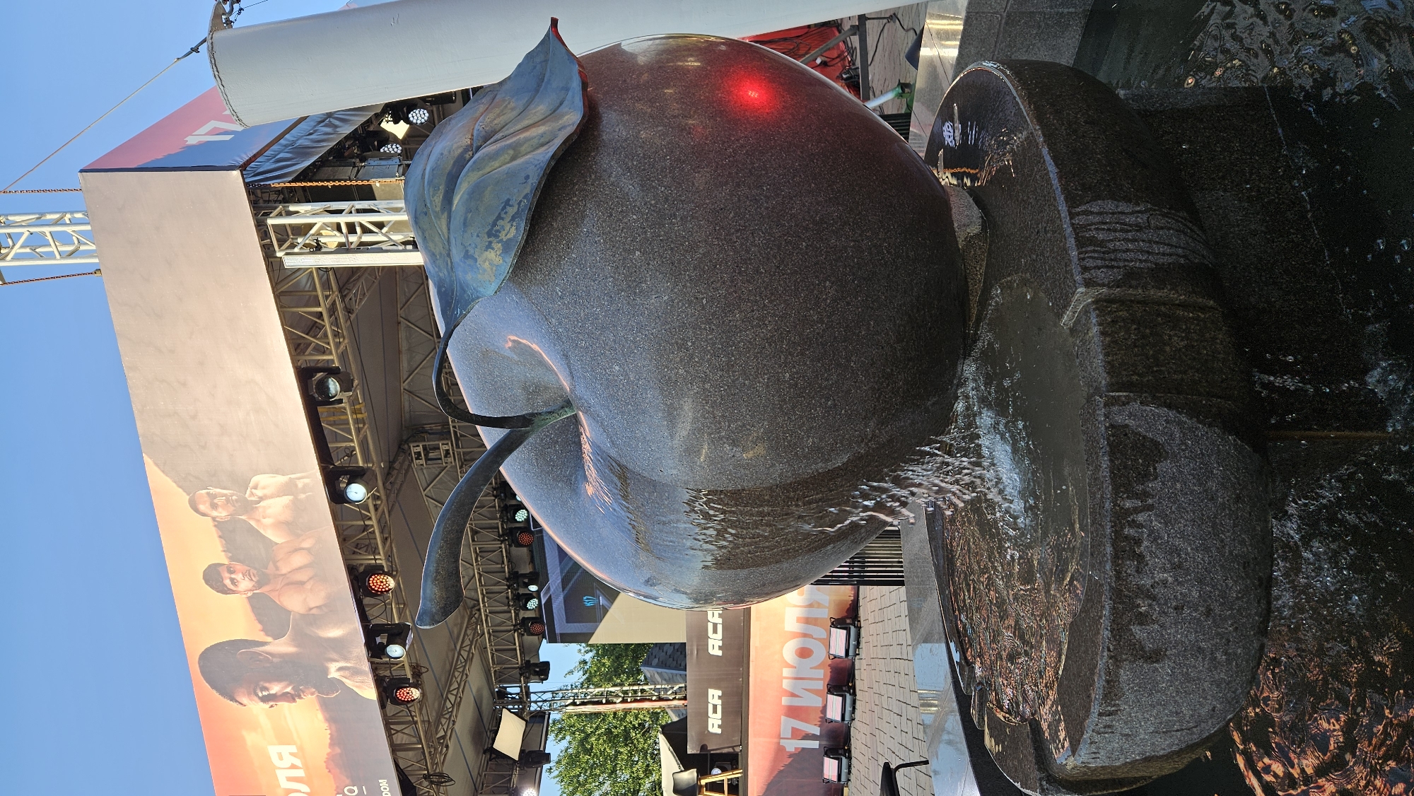 The image captures a close-up view of a granite apple fountain in the city of Almaty, Kazakhstan. The fountain is made of dark, speckled granite and features a large apple sculpture from which water cascades into a tiered basin. The water creates a dynamic effect with splashes and streams. A red light is visible on the side of the apple, possibly from a nearby event lighting. In the background, a large banner displaying images of three muscular men and text, including "17 ИЮЛЯ" (July 17th) and "ACA" (Absolute Championship Akhmat), is mounted above a stage structure with lighting equipment. The setting appears to be an outdoor venue, likely for a sporting event or festival, given the banner and stage. The sky above is a clear blue, suggesting it is daytime. The ground around the fountain is paved with light-colored tiles, reflecting the water and lights. The overall scene depicts a functional art piece within an event space.