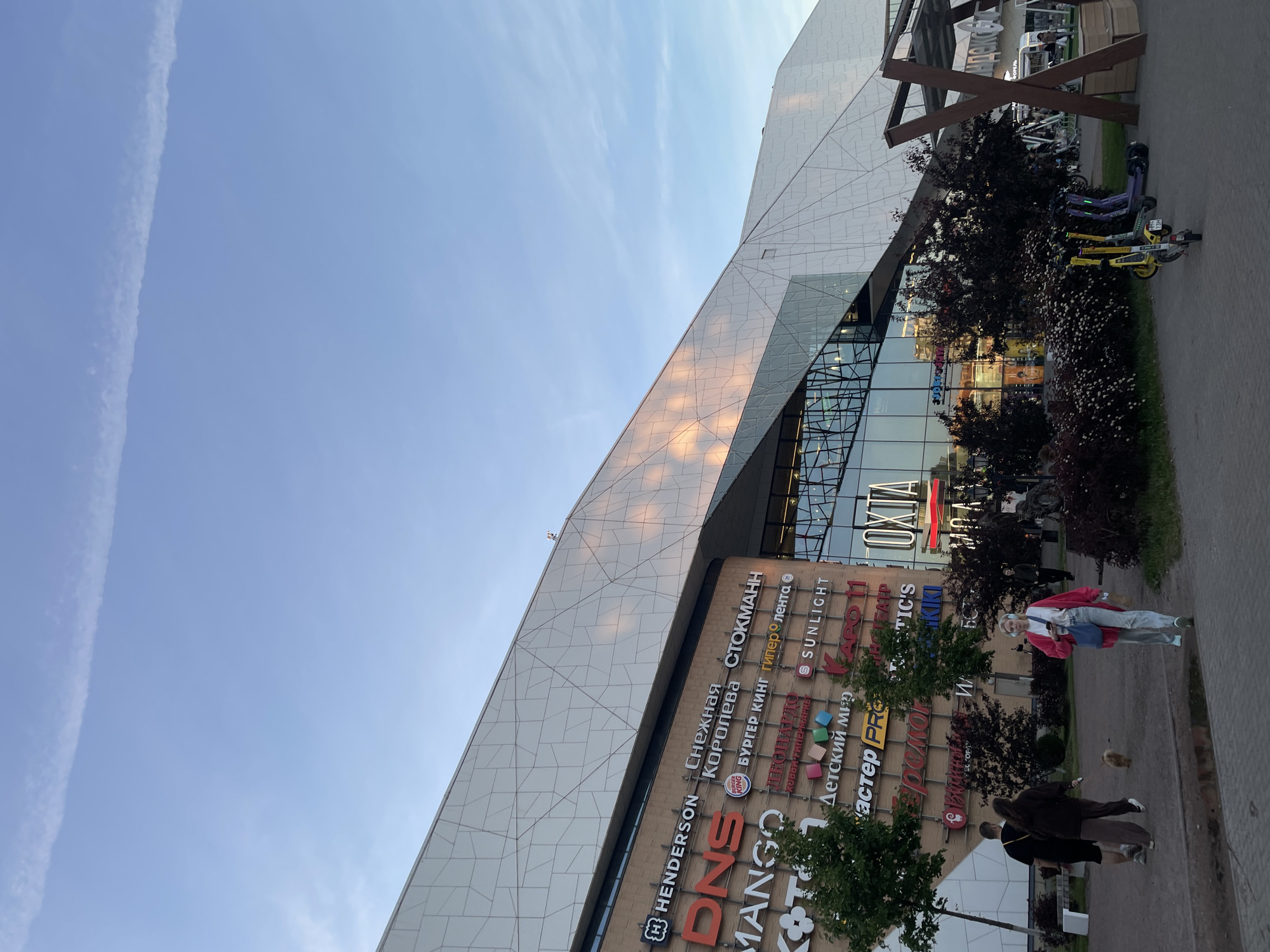 This image shows the exterior of the Okhta Mall, a shopping center located in the Krasnogvardeysky District of Saint Petersburg, Russia. The mall features a modern architectural design with a distinctive white, angular facade that reflects the light. Various storefront signs are visible on the lower brick portion of the building, including "HENDERSON," "DNS," "MANGO," "Снежная Королева," "СТОКМАНН," "BURGER KING," "Sunlight," and "ОХТА МОЛЛ."
In the foreground, several people are walking on a paved area. Two individuals, a man and a woman, are walking a small dog. A woman in a pink jacket and jeans is walking towards the camera, holding a phone and a paper bag. Several electric scooters are parked in a designated area to the right. Lush green trees and dark-leaved bushes surround the entrance area and the paved walkway. The sky above is a clear blue with wispy white clouds. The overall scene depicts a casual urban environment outside a commercial hub.