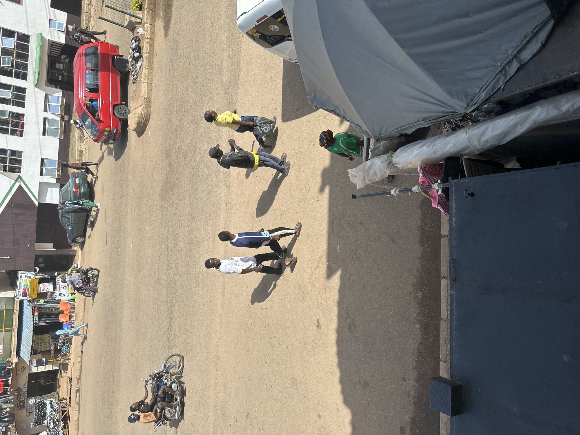 This is a high-angle, medium shot of a street scene in Bukuru, Nigeria. The street is paved with asphalt and appears to be moderately busy. In the foreground, to the left, is a dark, flat structure, possibly a building facade or a large object. To the right, a large, grey tarp or umbrella is positioned, obscuring parts of the scene.
In the middle ground, several people are walking or standing on the street. Two individuals are riding on a motorcycle, facing left. Further into the middle ground, four men are walking in a line from left to right, with shadows cast on the street. One man wears a white t-shirt and dark pants, the second wears a blue and white striped shirt and dark pants, the third wears a dark jacket and pants with a yellow shirt underneath, and the fourth wears a yellow t-shirt and dark pants, carrying a black bag. To the far right of this group, a woman in a yellow t-shirt walks away from the camera.
In the background, a red van is parked and occupied by a driver and a passenger. To the left of the van, a green car is also visible. Further back, several buildings line the street, with shops and awnings visible. A few individuals are seen near the buildings, including a person on a motorcycle. The overall environment suggests an urban setting with commercial activity.