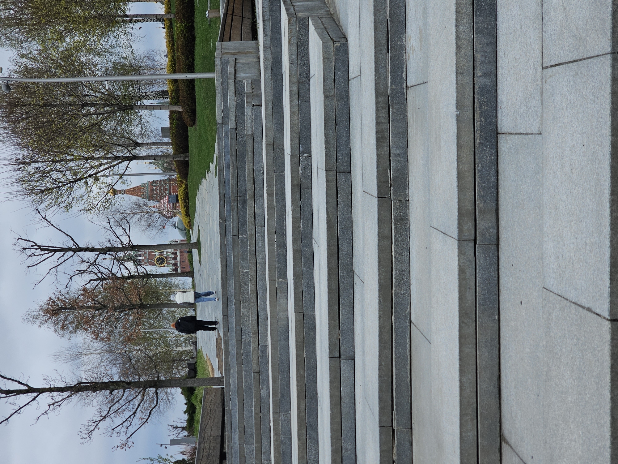The image depicts a set of stone stairs leading towards a view of the Moscow Kremlin, specifically the Spasskaya Tower. Two individuals are visible on the walkway at the top of the stairs. The trees in the background are bare, suggesting it is either early spring or late autumn. The stairs are constructed from grey stone slabs. Adjacent to the stairs is a grassy area and some bushes. The image is taken from a low angle, emphasizing the height of the stairs and the perspective towards the Kremlin. The sky is overcast.