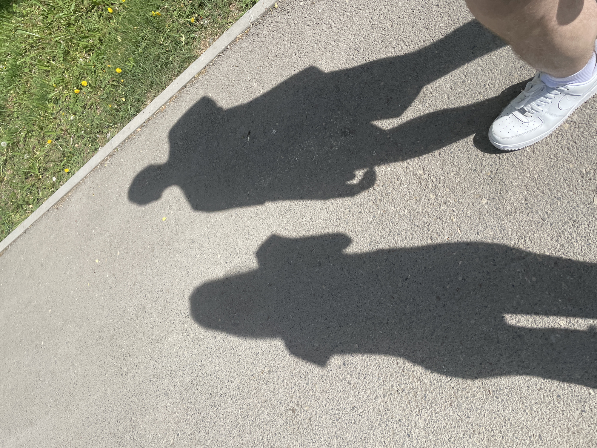 The image shows a concrete sidewalk adjacent to a patch of grass with yellow flowers. A person wearing white sneakers, shorts, and socks is standing on the sidewalk, casting a shadow. The shadow appears to be of the person holding a phone, based on the rectangular shape in the shadow. The location appears to be somewhere in Ufa, Russia.