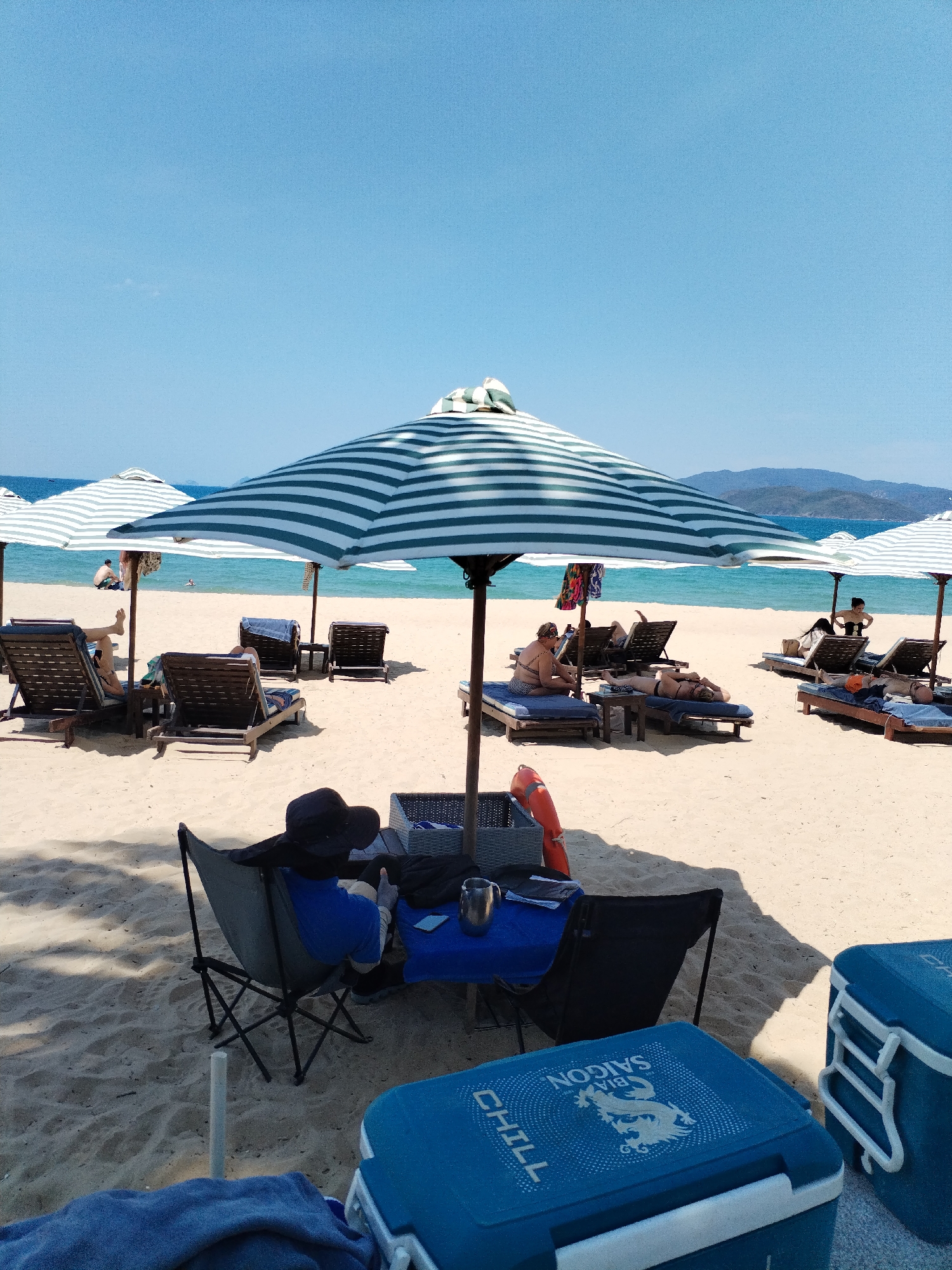 The image shows a beach scene with multiple beach umbrellas and lounge chairs on a sandy beach. People are relaxing on the lounge chairs. In the foreground, there are two blue coolers with logos and a small table with a person sitting behind it under an umbrella. The ocean and distant mountains are visible in the background under a clear blue sky. The location is Nha Trang Ward, Vietnam.