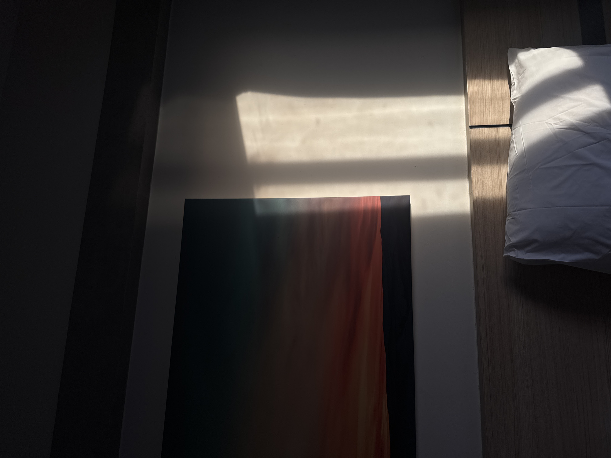 The image depicts a bedroom scene with a focus on a wall adorned with a landscape artwork and illuminated by dramatic natural light casting shadows. The artwork, a rectangular canvas, displays a gradient of colors transitioning from deep teal at the top to warm reds and oranges, below which is a dark, silhouetted landscape of hills. To the right of the artwork, a wall is illuminated by shafts of light, creating distinct rectangular and elongated shapes of brightness against a darker background. The lighting suggests a low sun angle, likely from a window. Below the artwork and the illuminated wall, the top portion of a wooden headboard is visible, with a white pillow resting on it. The pillow is partially lit by the same natural light, showing soft folds and shadows. A thin, dark vertical object, possibly a book or a remote, is subtly placed behind the pillow. The room appears to be a bedroom, possibly in a hotel or residence, in Surat Thani City Municipality, Thailand. The overall mood is serene and atmospheric due to the interplay of light and shadow.