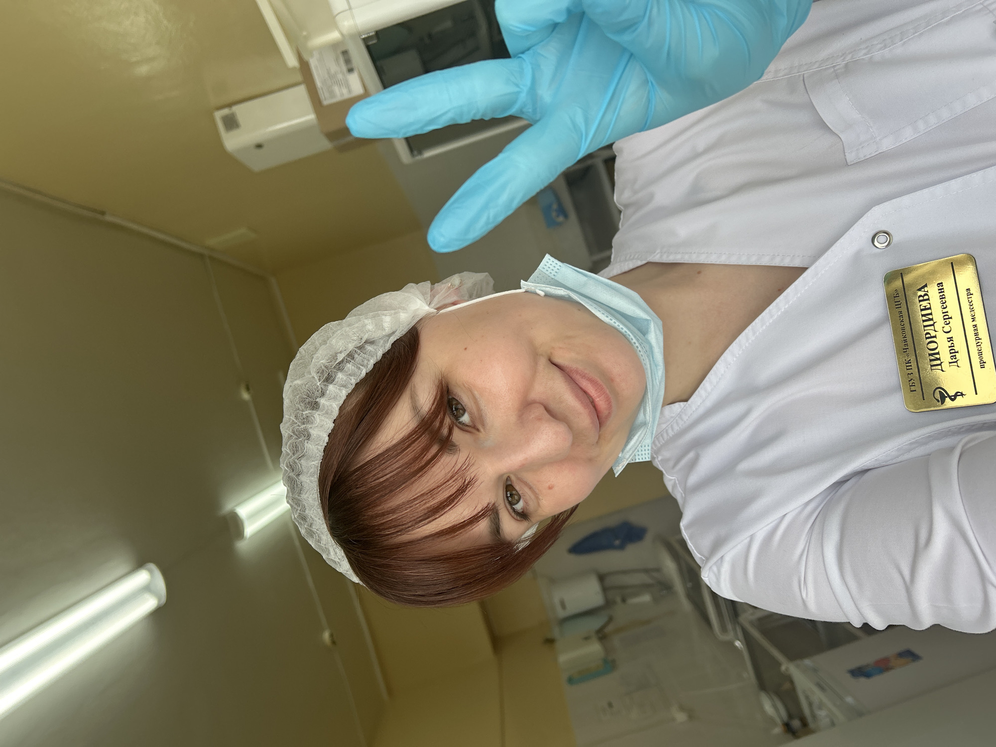 A cheerful selfie shows a woman, identified as Darya Sergeevna Diordieva, a procedure nurse, in what appears to be a medical setting in Fokino, Russia. She is wearing a white medical coat, a disposable white hairnet, and a light blue medical mask pulled down to her chin. Her reddish-brown hair is visible beneath the hairnet. She has brown eyes and is smiling faintly, looking directly into the camera. Her left hand, encased in a blue medical glove, is raised, forming a 'V' or peace sign.
The background suggests an indoor medical facility, with light-colored (possibly beige or light yellow) walls and ceiling. Fluorescent strip lights are visible on the ceiling, illuminating the room. Other indistinct medical equipment or shelving can be seen in the background.
A name tag pinned to her white coat is clearly visible and reads in Russian:
* ГБУЗ ПК «Ханкайская ЦРБ» (State Budgetary Healthcare Institution of Primorsky Krai "Khankayskaya Central District Hospital")
* ДИОРДИЕВА (DIORDIEVA)
* Дарья Сергеевна (Darya Sergeevna)
* процедурная медсестра (procedure nurse)