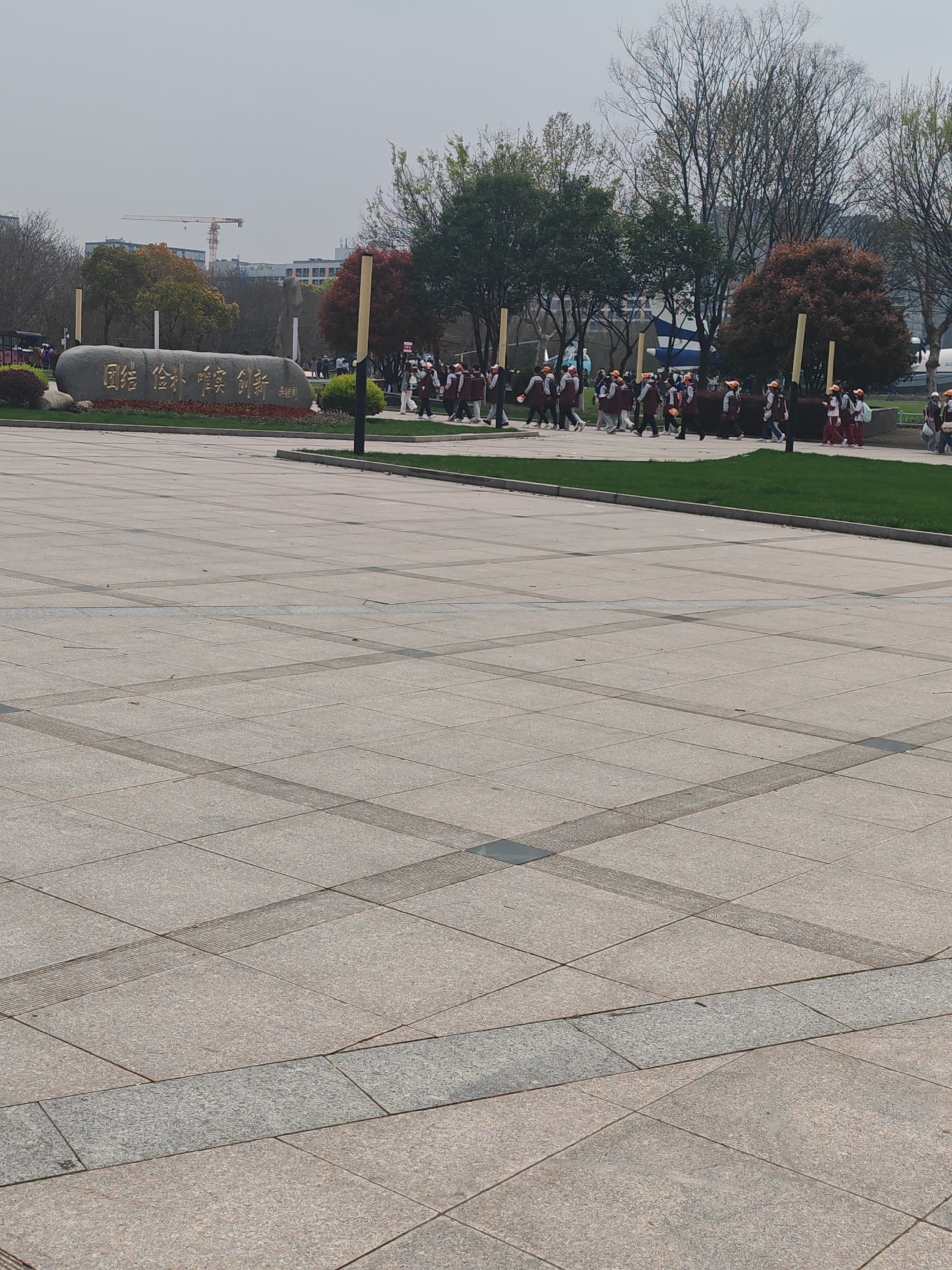 The image shows a large paved plaza with a group of people walking along a grassy strip. To the left is a stone monument with Chinese characters on it. In the background, there are trees, buildings, and a construction crane. The location is the city of Jiangning District, China, based on the Chinese characters on the stone monument. The group of people are wearing similar clothing, suggesting they are together for an organized activity. The weather appears to be overcast.