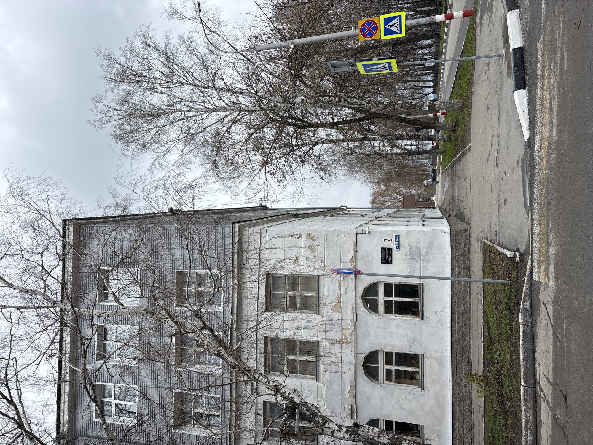 The image captures a quiet street scene in an urban or suburban setting, likely in Russia during late autumn or early winter. The sky is overcast and grey, casting a subdued light over the area.
On the left, a multi-story building stands, partially framed by the dense, bare branches of a large tree in the foreground. The building's facade is sectioned: the left portion is a grey, possibly corrugated or paneled material with rectangular windows, while the right portion, facing the street, is lighter in color (white or light grey) with visible peeling paint or stucco. This lighter section features a mix of rectangular and distinctive arched windows. A dark plaque is mounted on this part of the building, clearly showing the number "2" and the Cyrillic text "ул.КОРОБИХИНА," indicating the street name "KOROBIKHINA street."
To the right of the building, a concrete sidewalk runs alongside an asphalt road. A black and white striped curb marks the edge of the road. Sparse green grass can be seen in patches along the sidewalk, where more bare deciduous trees line the street into the distance.
Above the trees, a pole extends over the road, supporting several street signs. Visible signs include two blue square pedestrian crossing signs (featuring a white triangle with a walking person) and one circular "no stopping" or "no parking" sign (blue with a red diagonal line and border). Another tall pole, possibly holding a streetlight or camera, is visible further up.
There are no people or active events taking place, contributing to the tranquil and somewhat stark mood of the scene. The bare trees and overcast weather suggest a cold, possibly pre-winter day.
