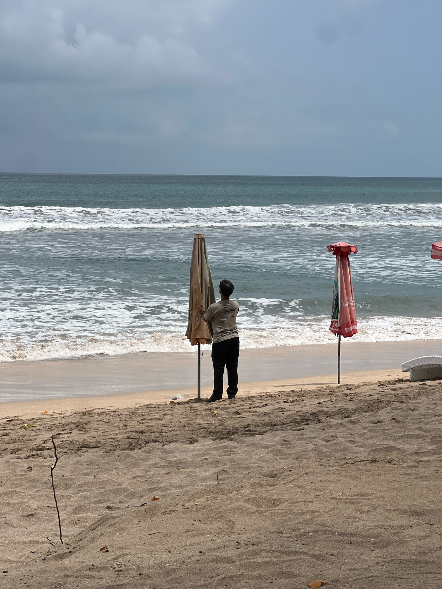 A person is standing on a sandy beach in Kuta, Indonesia, facing away from the camera and toward the ocean. The person, wearing dark pants and a light-colored t-shirt, is near a closed, tan-colored beach umbrella. To the right of this umbrella, another closed beach umbrella with red and white fabric is visible. The ocean is in the background with moderate waves breaking on the shore. The sky is overcast with gray clouds. The sand in the foreground is textured with small debris and dried leaves. In the far right of the image, a white lounge chair is partially visible.