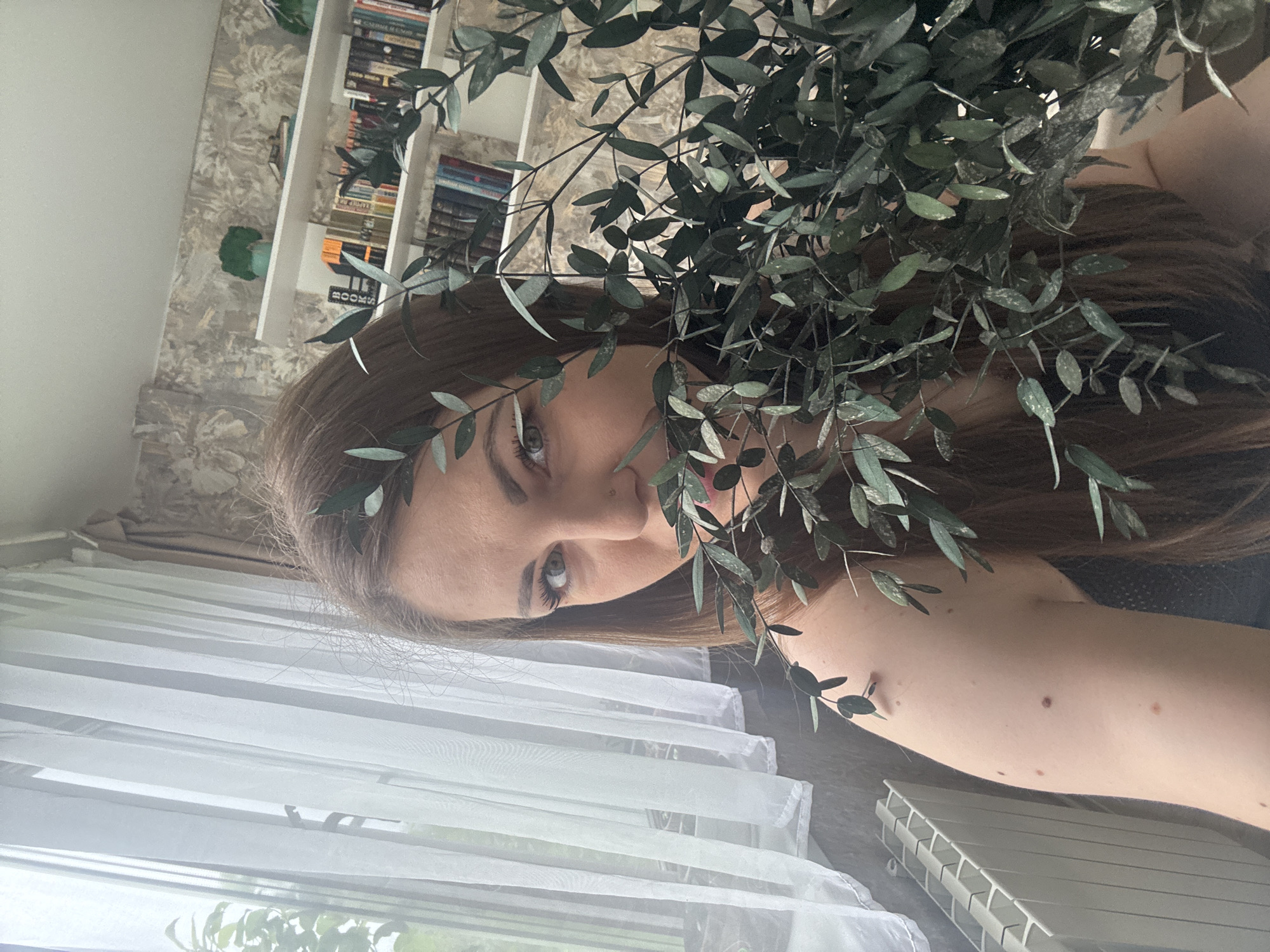 A woman with long brown hair is holding a leafy green branch in front of her face. She has fair skin and is wearing a dark-colored top. The background includes a bookshelf with books and a window with sheer white curtains. The image appears to be taken indoors. It's not possible to determine whether this is in the city of Раздолье, Russia.
