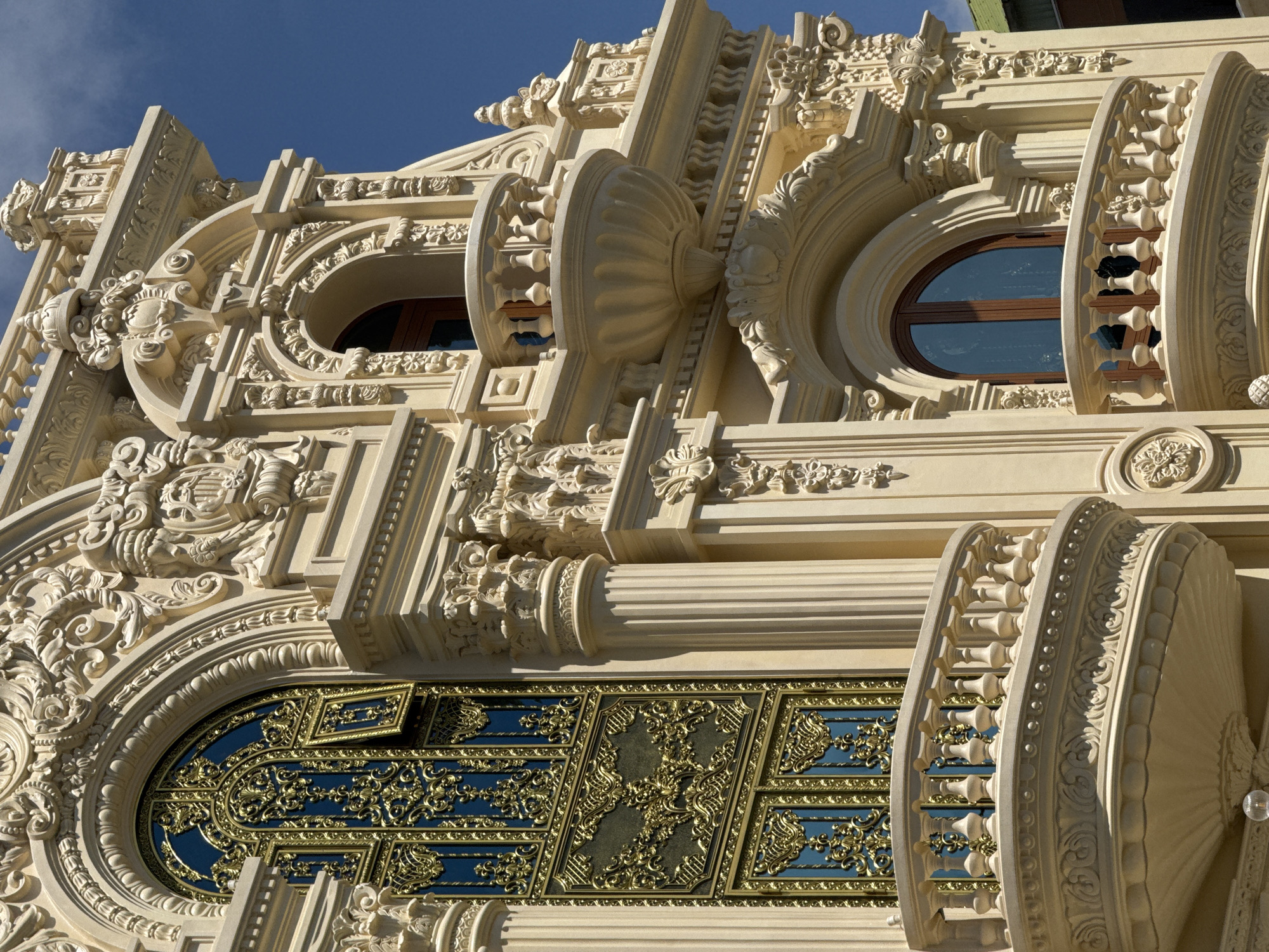 The image shows the ornate exterior of a building in Da Lat, Vietnam. The architecture is characterized by elaborate decorative elements, including intricate carvings, columns, and balconies. A large window with a golden, filigree-like grille and blue panes is prominently featured on the left side of the building. Several balconies with decorative railings are visible, jutting out from the facade. The building is painted in a pale, cream-like color and stands against a clear blue sky. No people are visible in the frame. The overall impression is one of opulence and intricate craftsmanship.