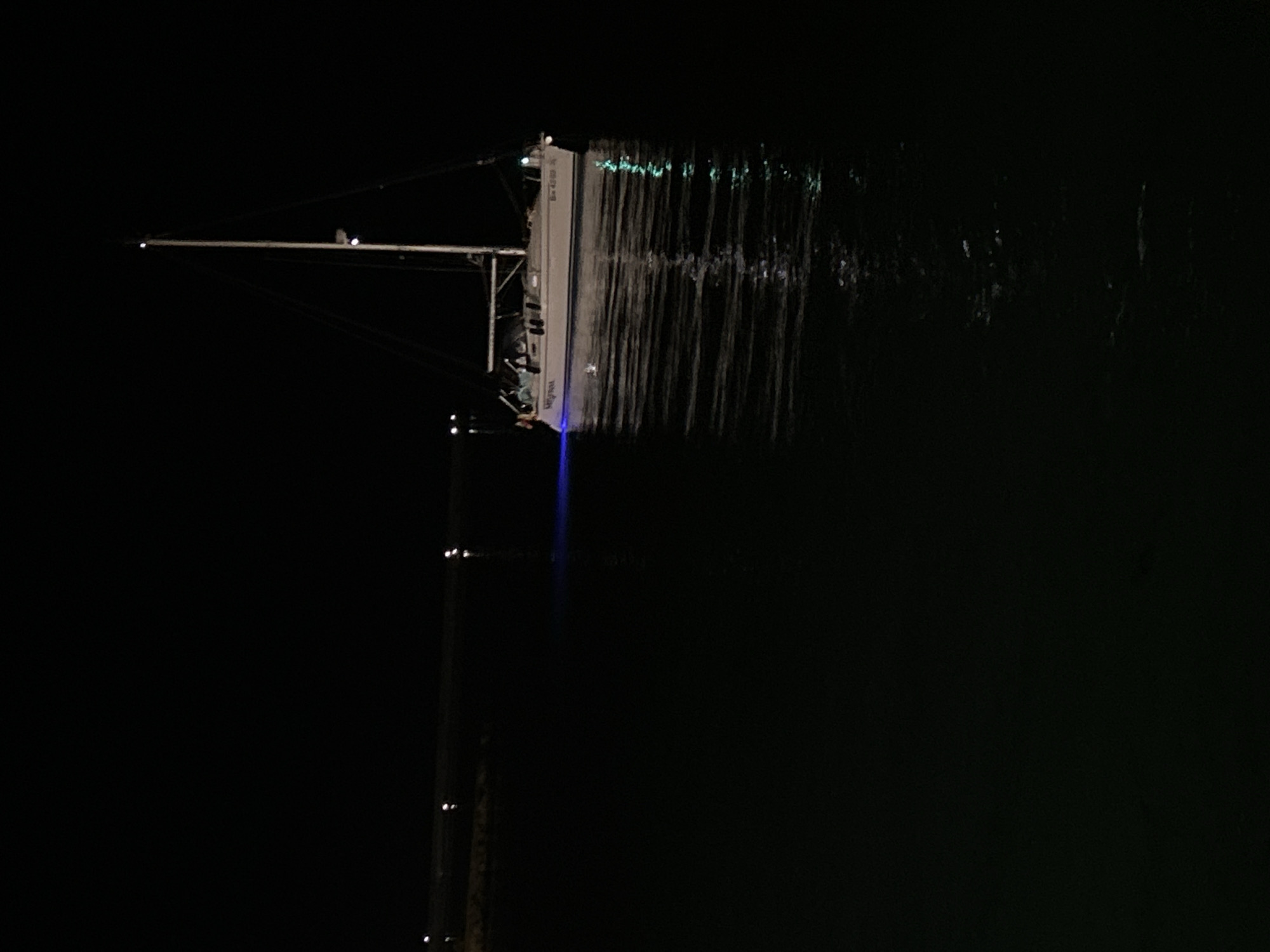 The image depicts a nighttime scene in Balchik, Bulgaria, featuring a single white-hulled monohull sailboat floating on calm dark water. The sky is completely black, indicating a lack of natural light.
The sailboat is positioned centrally in the frame. Its mast extends vertically upwards, equipped with various rigging lines. A light, likely white, is visible near the top of the mast. On the starboard side of the boat (viewer's left), a green navigation light is illuminated, casting a green reflection onto the water. Towards the stern, an underwater or transom light emits a distinct blue glow into the water immediately behind and to the side of the vessel, creating a clear blue luminescence on the surface. The name "Myrina" is visible on the hull, along with a registration number "Bx 4369 K" on the stern. No individuals are clearly discernible on board the boat.
The water surface exhibits gentle ripples, reflecting the boat's blue and green lights, as well as several smaller, indistinct white reflections. In the distant background, along the horizon on the left side, a dark landmass or structure, possibly a pier or coastline, is visible, marked by several small, faint white lights that also produce subtle reflections on the water. The overall environment is dark and serene, illuminated solely by artificial light sources.