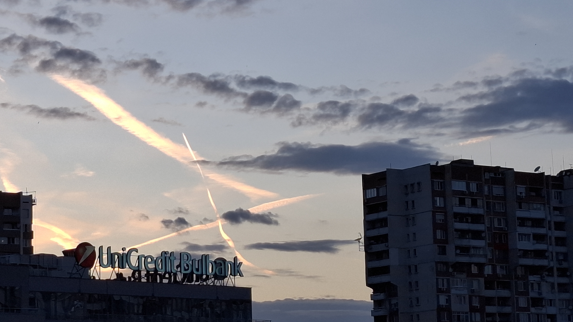 The image shows a twilight scene in Sofia, Bulgaria. The sky features clouds and prominent contrails from aircraft. In the foreground is the Unicredit Bulbank sign on top of a building. To the right is a multi-story residential building.