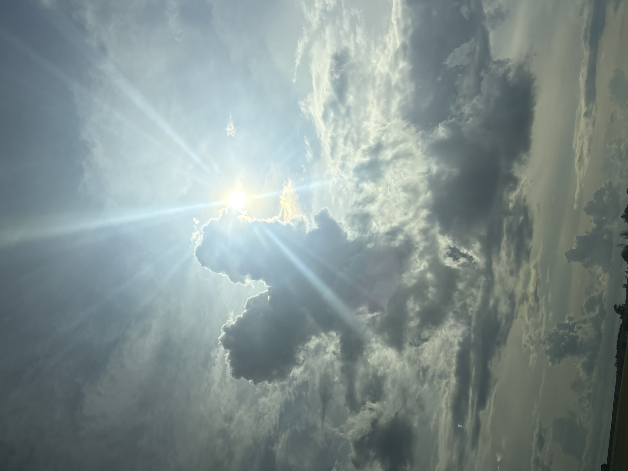 This is a photograph of a sky filled with dramatic clouds and sunbeams. The sun is partially obscured by a large, dark cloud formation in the upper left quadrant, with bright rays of light emanating outwards. The sky is a mixture of light blue and gray, with varying densities of clouds. The lower portion of the image shows a sliver of a landscape with a field and trees, suggesting an outdoor setting. The location is specified as Сеніцкі сельскі Савет, Belarus. There are no people or specific objects visible that would indicate an action or interaction. The scene is a natural landscape under a dynamic sky.