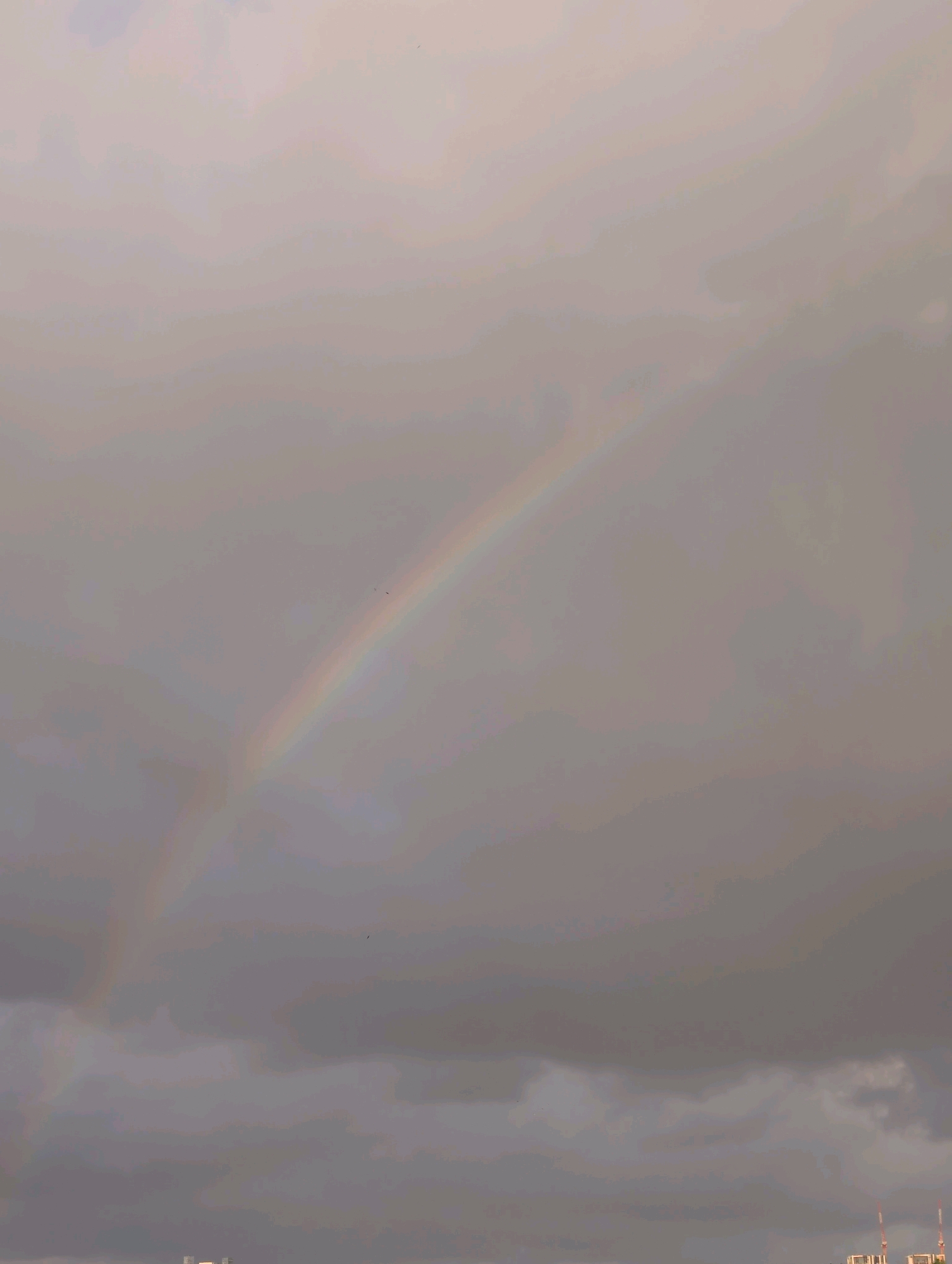 A double rainbow arcs across a sky filled with dense, grey clouds. The faint colors of the rainbow are visible against the muted backdrop. In the distance, the indistinct silhouettes of buildings and structures, part of the cityscape of Ramat Gan, Israel, are visible along the horizon.