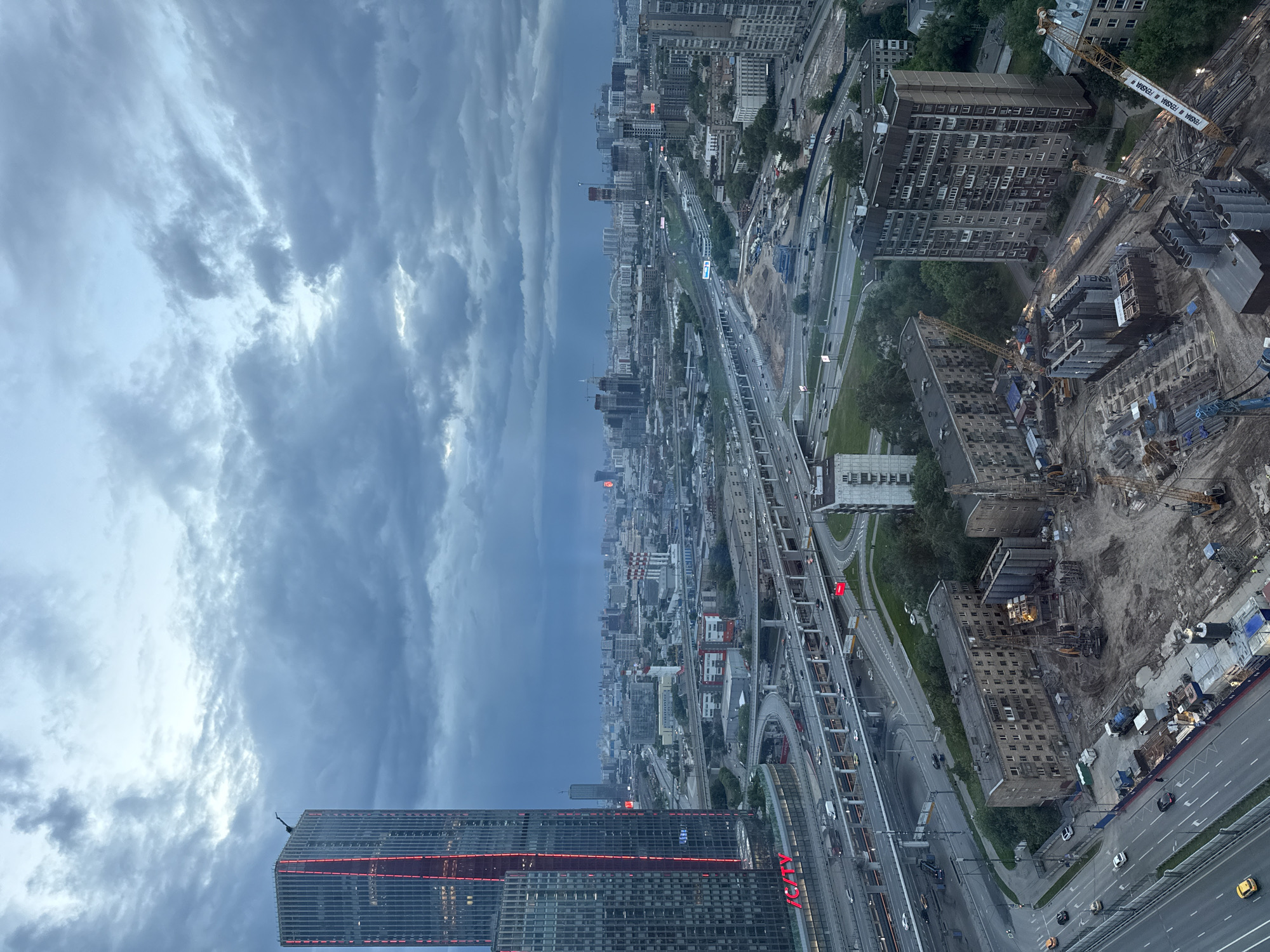 The image shows an aerial view of a city, likely Khimki, Russia, under a cloudy sky. In the foreground, a modern skyscraper with red accents is visible. The city stretches into the distance, with numerous buildings of varying heights, including both residential and commercial structures. A complex network of roads and overpasses crisscrosses the urban landscape, with visible traffic. Construction sites are present, indicated by the presence of cranes and unfinished buildings. The overall environment appears to be urban, dense, and undergoing construction and development.