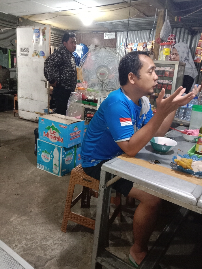 The image depicts an indoor scene within a casual eatery or small shop in Kumpulrejo, Indonesia. Three individuals are visible. In the foreground, a man in a blue and white short-sleeved shirt with an Indonesian flag emblem is seated on a brown plastic woven stool at a grey metal-framed table. He faces right in profile, with both hands raised and open, gesturing as if speaking. On the table in front of him are a light blue bowl, a white plate with fried food items, and several plastic bottles. His feet, wearing light green flip-flops, are visible beneath the table.

Behind him, to the left, a man in a black and white patterned long-sleeved jacket and dark pants stands with his hands in his pockets, looking towards the right. To the man in the blue shirt's right and slightly behind him, a woman wearing a grey hijab is partially visible behind a display counter containing various packaged goods. A large white wall-mounted electric fan is positioned between the two men.

The environment features a concrete floor and what appears to be a corrugated metal ceiling or roof section. To the left, a white wooden or painted panel with the word "KASIR" (Cashier) written on it is visible. Stacked on the floor near the cashier panel are two blue cardboard boxes of "Le Minerale" bottled water. Shelves displaying various products line the wall behind the woman. The scene is illuminated by overhead artificial lighting.