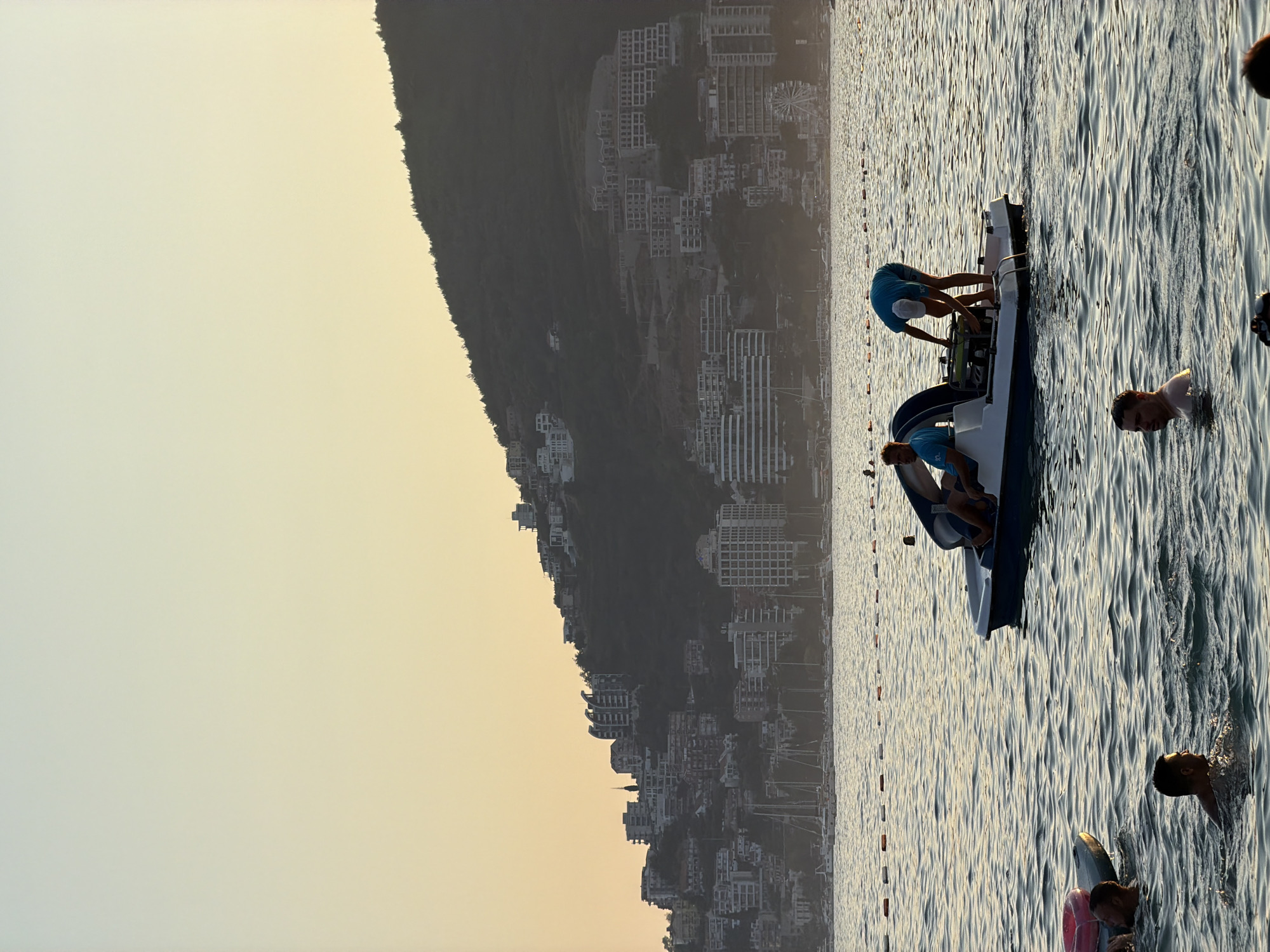 The image depicts a serene coastal scene in Bečići, Montenegro, during what appears to be late afternoon or early evening, as indicated by the soft, golden light and the gradient of the sky from pale yellow to light blue. In the foreground, several individuals are visible in the calm, rippling water. One man is swimming, looking towards the right side of the frame. To his left, another man is partially submerged, with his head and shoulders above the water, and he appears to be near an inflatable object. Further to the left, another person is visible in the water.
The middle ground features a pedal boat with two individuals. One person is seated, and another is standing at the back of the boat, appearing to adjust or operate some equipment. A line of buoys stretches across the water, marking a boundary or swimming area.
In the background, a hillside is densely covered with buildings, ranging from smaller structures to multi-story apartment complexes and hotels, creating a picturesque urban landscape cascading down towards the sea. Sailboats are anchored or moored in the bay below the hillside. The overall atmosphere is tranquil, with the gentle lapping of waves and the warm glow of the setting sun.