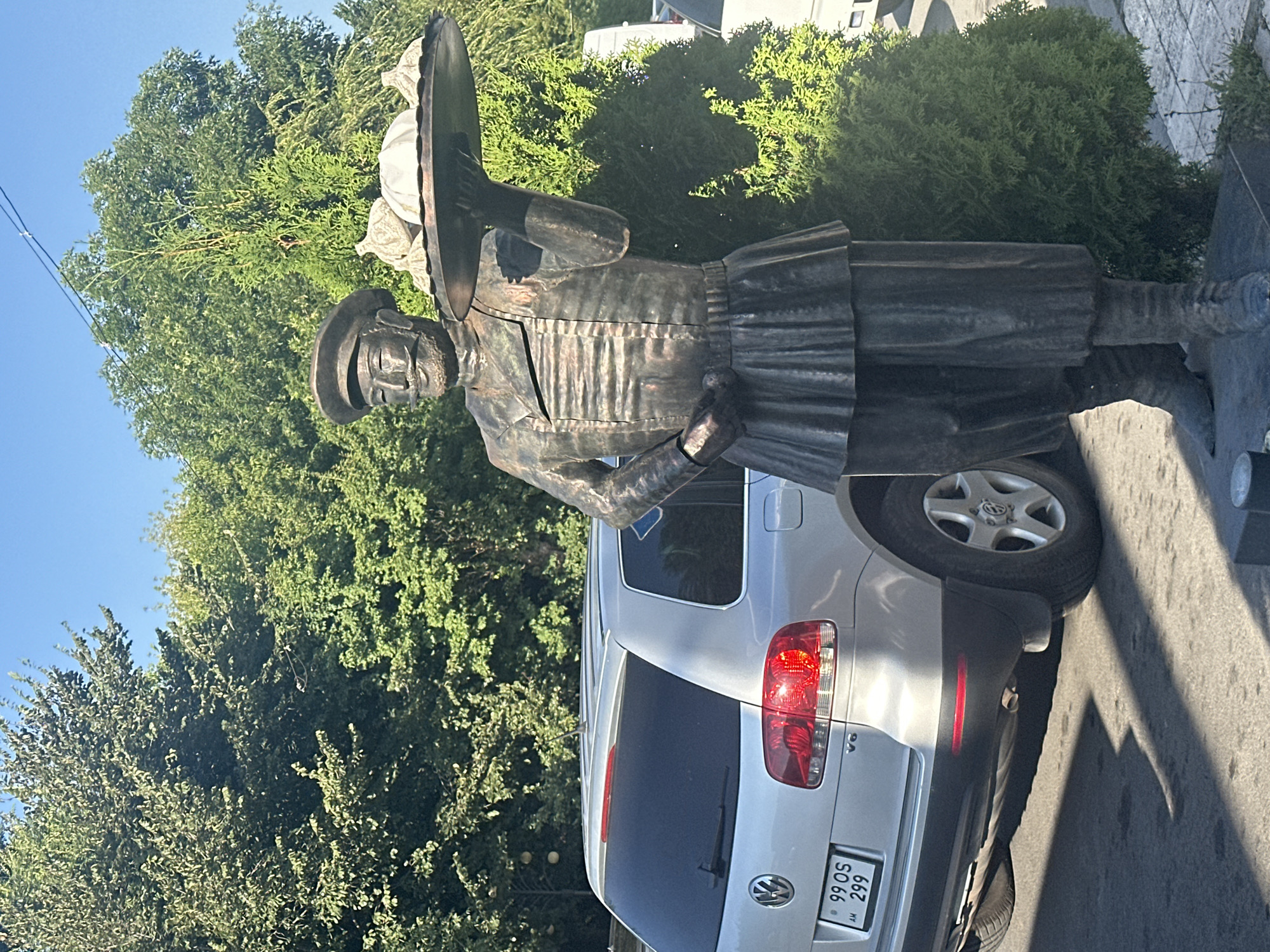 The image shows a bronze statue of a man in traditional attire, holding a tray with food. The statue is located in Gyumri, Armenia, likely in a public space. The man is depicted wearing a cap, a long-sleeved jacket with a pleated collar, and a skirt-like garment over pants. His facial features include a mustache and a beard. He is holding a circular tray with several items on it, possibly food items like khinkali (dumplings) and bread. Behind the statue, a silver Volkswagen Touareg SUV is parked. The environment appears to be outdoors, with lush green trees and a clear blue sky. The ground is paved with asphalt. The license plate of the car reads "99 OS 299 AM". The statue is positioned on a pedestal.