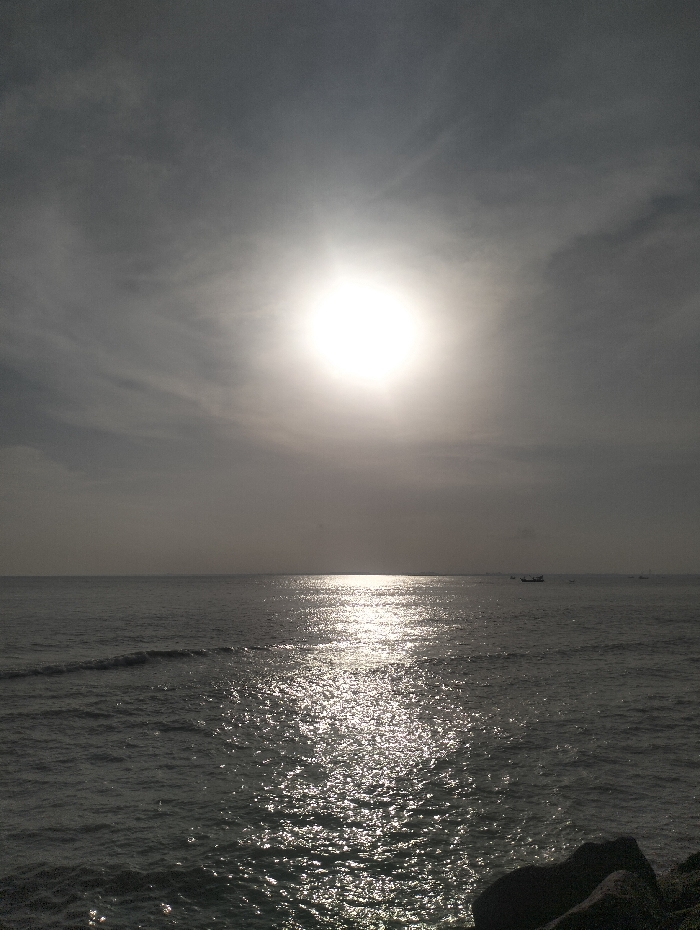 A wide, glistening ocean stretches to the horizon under a hazy sky. The sun, high in the sky, blazes intensely, its light reflected in shimmering patterns across the water's surface. Small waves ripple towards the foreground. In the distance, a tiny boat is faintly visible near the horizon. Dark rocks occupy the bottom right corner of the frame. The overall scene is monochromatic and luminous.