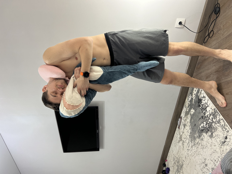 An adult Caucasian male, shirtless and wearing dark grey athletic shorts, stands barefoot indoors in a room in Kazan, Russia. He has a light pink U-shaped neck pillow around his neck. He is cradling a large, light blue and beige shark plush toy against his upper body, with his closed eyes and head resting on the toy's head, suggesting a state of sleep or deep relaxation. An orange-banded smartwatch is present on his left wrist.
The background consists of a plain white wall with a dark, wall-mounted Samsung flat-screen television positioned to the subject's left. The floor features dark wood laminate, partially covered by a light-colored rug exhibiting abstract grey and black patterns. In the lower left corner, on the rug, several small, dark spherical objects and a purple soft item are visible. To the subject's right, a white electrical outlet is visible with a black cord plugged into it, featuring a foot-operated switch resting on the floor.