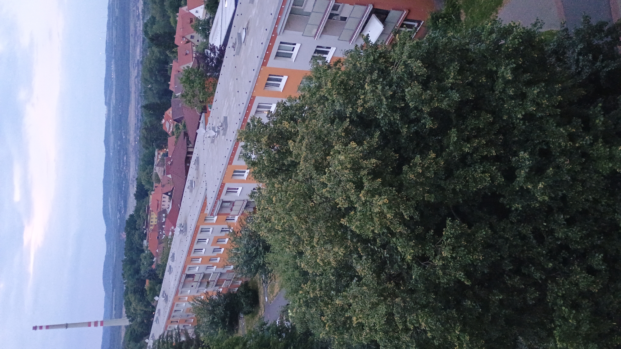 The image depicts a view of a residential area in Sokolov, Czechia, with a large apartment building in the foreground. The apartment building is multi-storied and features a light gray facade with orange accents around the windows and balconies. The balconies have railings and some appear to have awnings. The roof of the building is flat and gray, with several satellite dishes and ventilation structures visible.
In front of the apartment building, a large, dense green tree with abundant foliage dominates the lower half of the image, obscuring much of the ground level. To the left of the apartment building, a paved walkway or road is visible, bordered by grass.
In the background, beyond the apartment building, a panorama of a town or city is visible. This includes numerous residential houses with red-tiled roofs, interspersed with trees. Further in the distance, rolling hills and mountains form the horizon under a cloudy, overcast sky. A prominent, tall, industrial chimney with red and white stripes stands on the left side of the distant landscape, possibly indicating industrial activity in the vicinity. Wind turbines are also faintly visible on a distant hill. The overall impression is a daytime scene in a moderately built-up area, likely in the late afternoon or early evening given the muted lighting.