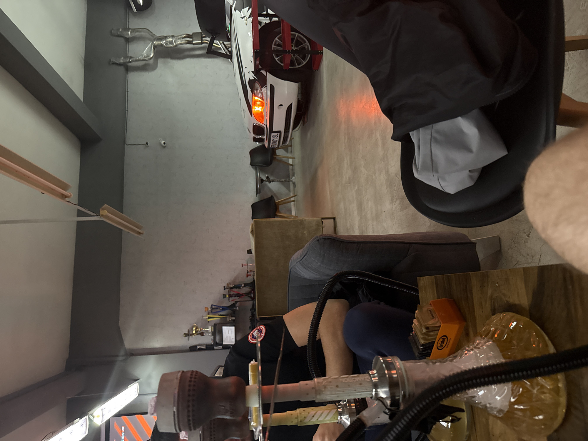 A hookah is positioned in the foreground of an indoor space. In the background, a vehicle is in what appears to be a repair bay. Trophies are displayed on a shelf in the background. A person wearing a black shirt with a round logo on the shoulder is seated near the hookah. A lighter rests on a small wooden table next to the hookah. The exact location within Plovdiv, Bulgaria, cannot be determined from the image.