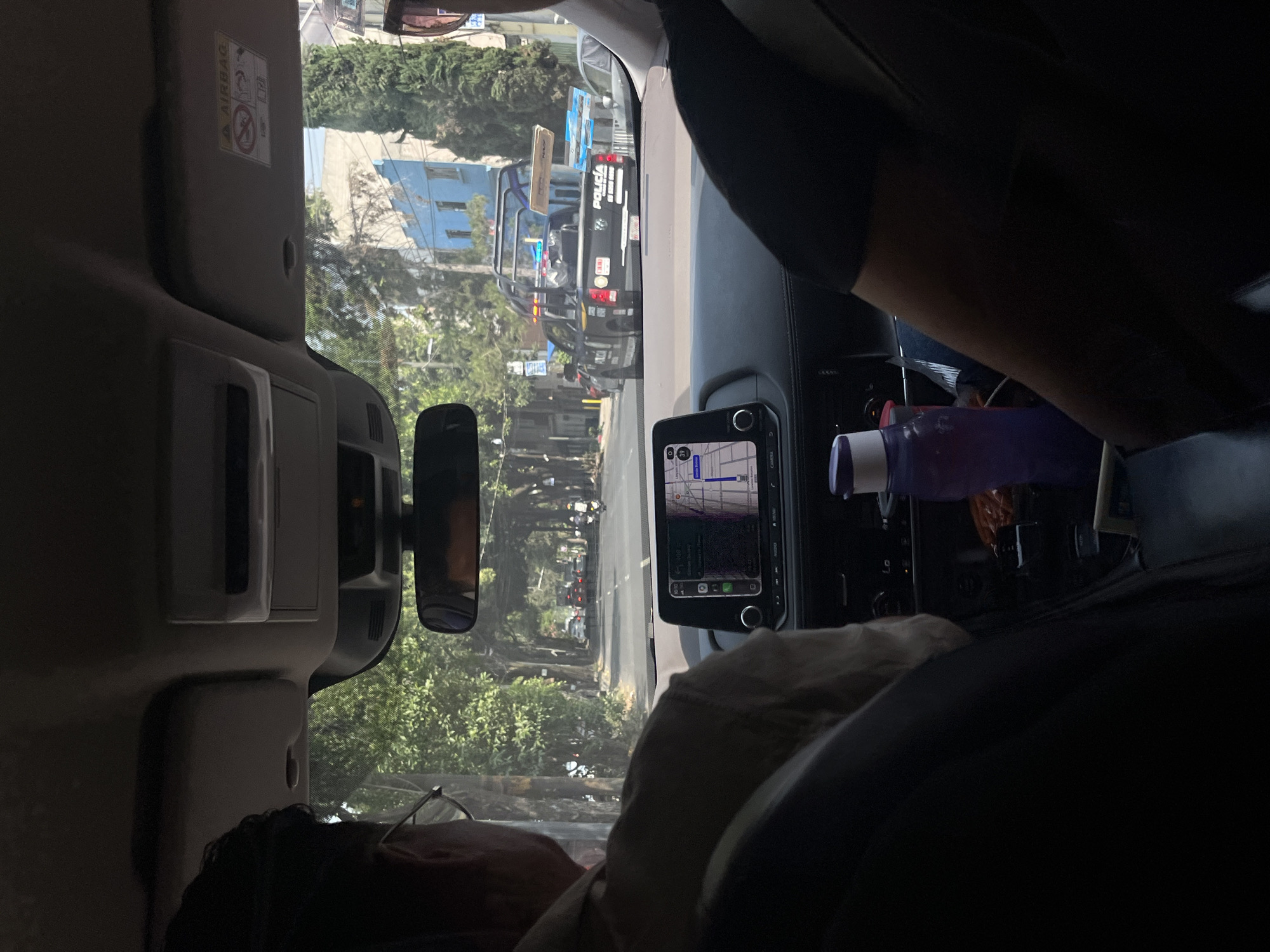 A view from inside a vehicle in Mexico City shows a street lined with trees and buildings. In the foreground, a person with glasses is partially visible in the lower left. The vehicle's dashboard is partially visible, featuring a navigation screen displaying a map. A water bottle is present on the dashboard. Through the windshield, a police vehicle is visible on the street ahead. The scene suggests a typical urban environment with law enforcement presence.
