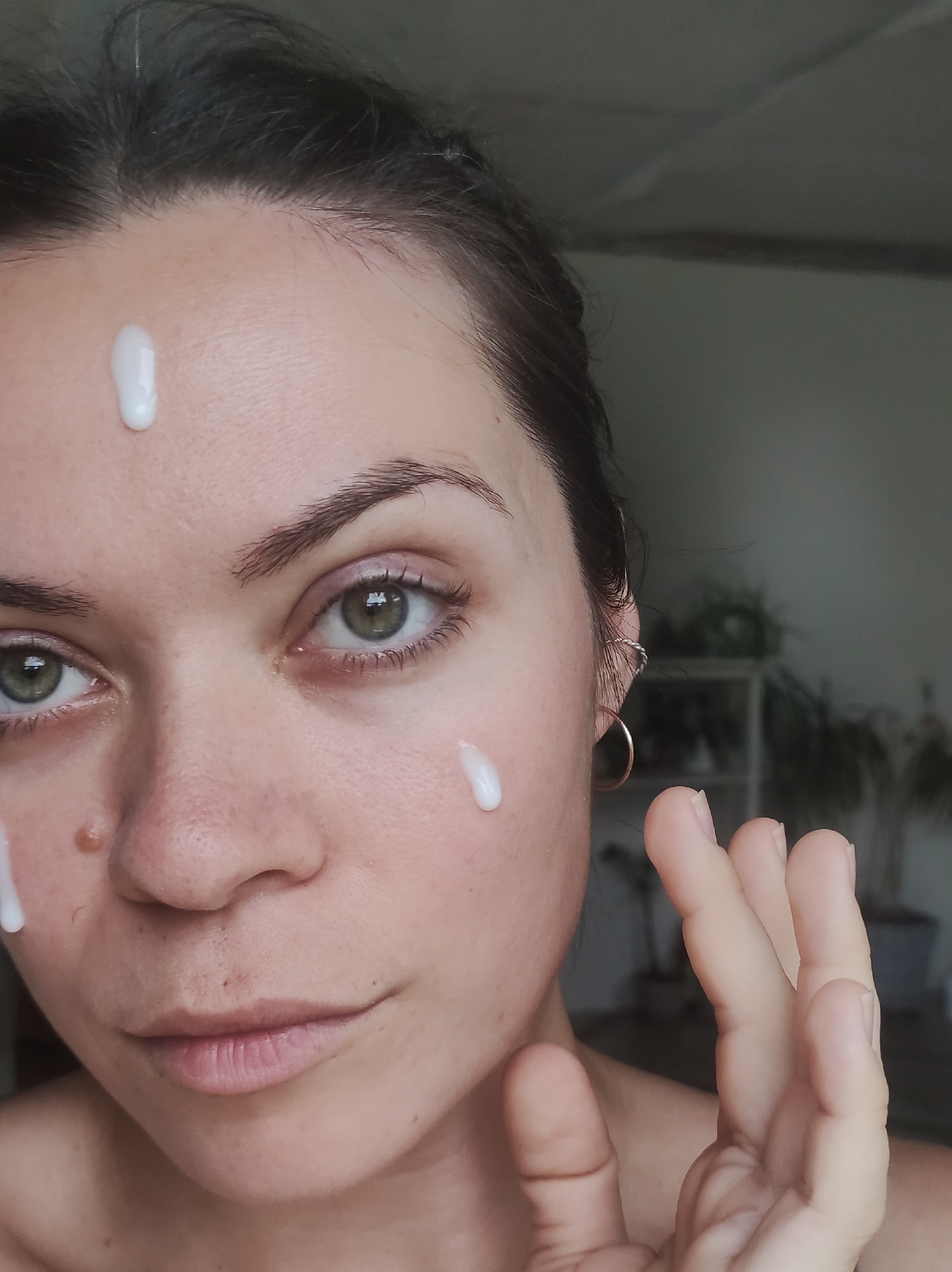 A close-up, indoor shot captures a woman from the chest up, applying a white, viscous substance to her face. Her head is slightly tilted, and her gaze is directed towards the viewer with a neutral expression. Three distinct droplets of the white product are visible: one centrally on her forehead, one on her right cheek, and a third partially visible below a prominent brown mole on her left cheek.
The woman has dark hair, pulled back, revealing her forehead and ears. Her eyes are open and appear green. Her eyebrows are naturally shaped. Her lips are closed, displaying a natural, uncolored appearance. On her right ear, a thin, gold-colored hoop earring is visible, along with a smaller, twisted silver-colored earring higher on the earlobe. Her left hand is raised to the right side of her face (viewer's right), fingers slightly splayed, poised as if to spread the product. Her nails are short and unpolished.
The background is softly blurred, indicating an indoor setting with shallow depth of field. A neutral grey wall is visible behind her. To the right, a faint, light-colored vertical structure, possibly a shelf or part of furniture, holds dark green leafy plants. The overall lighting is soft and even. The specific location in the city of Фоки, Russia, is not determinable from the visual information provided in the image.
