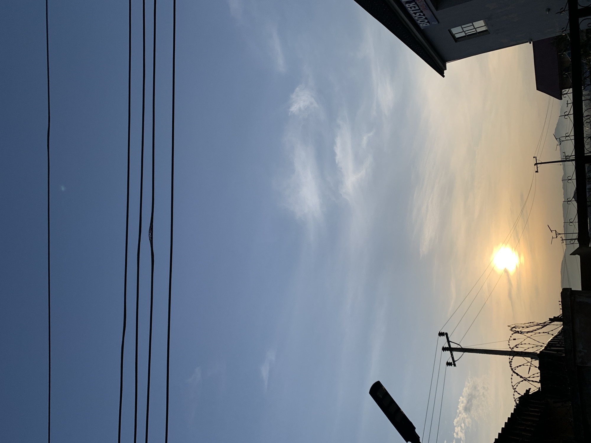 The image captures a cityscape during what appears to be sunset or sunrise in Rafin Par, Nigeria. The sky is a gradient of deep blue at the top, transitioning to lighter blue and then a warm yellow-orange hue near the horizon. Several black power lines are stretched across the upper portion of the frame, with some exhibiting slight sagging. A street lamp is silhouetted on the left side of the image. In the middle ground, a power pole with insulators supports more power lines. To the left, a wall topped with barbed wire fencing is visible. In the foreground and right side of the image, the corner of a building is shown, with a sign that reads "BOOSTER". The building is a light color with a dark roof and a window. In the distance, a hazy mountainous landscape is discernible. The overall scene is one of urban infrastructure set against a natural sky.