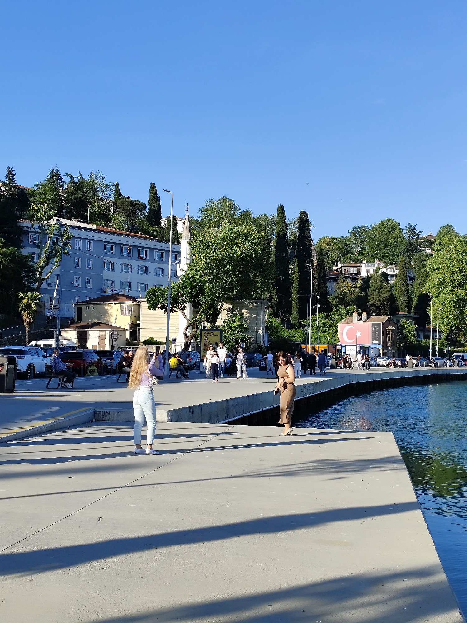 The image depicts a waterfront scene in Sarıyer, Turkey, on a sunny day. Two women stand on a concrete pier next to the water. The woman on the left wears a purple top and light jeans and appears to be taking a picture. The woman on the right wears a tan dress and stands near the water's edge. Behind them, a crowd of people stroll along the waterfront, with parked cars visible in the background. Buildings and trees line the hillside in the background. Notably, there is a building decorated with the Turkish flag. A mosque is also visible in the background. The pier casts long shadows on the concrete.