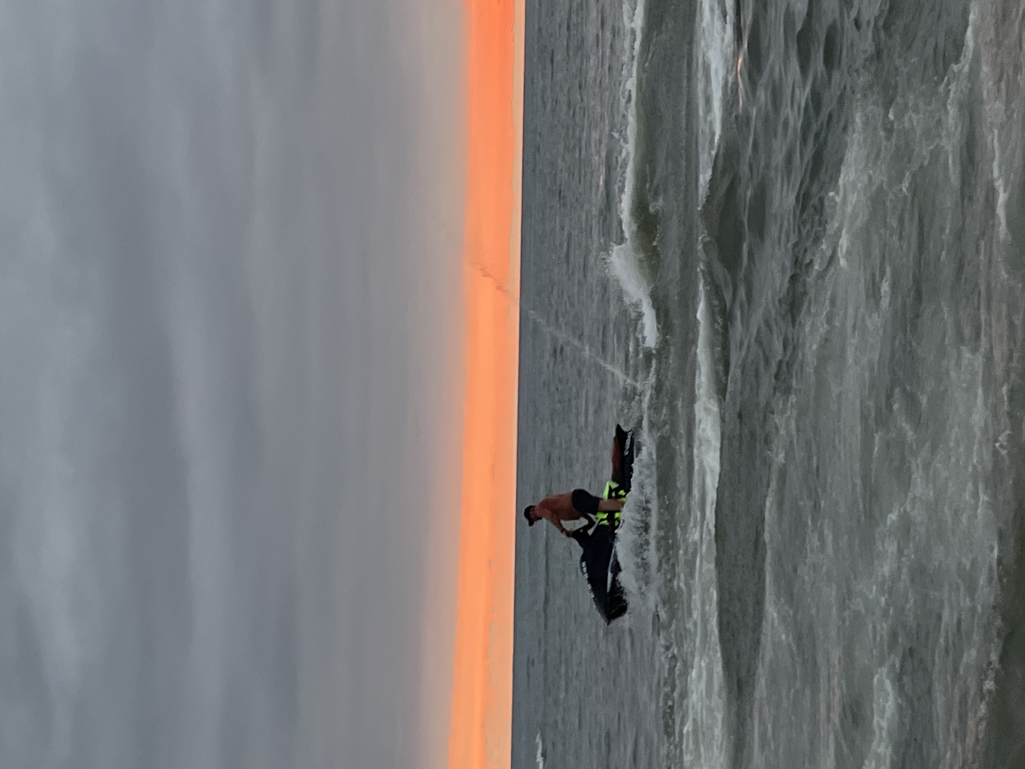 A person rides a black jet ski in choppy gray water under a sky with a vibrant orange band near the horizon. The person is wearing a reddish-orange shirt, dark shorts, and a dark baseball cap. A bright yellow life vest is visible. The jet ski is creating a wake with white foam. In the background, the ocean stretches to the horizon, where the bright orange band of sunset meets the darker gray clouds. The location is the Black Sea coast in Ureki, Georgia.
