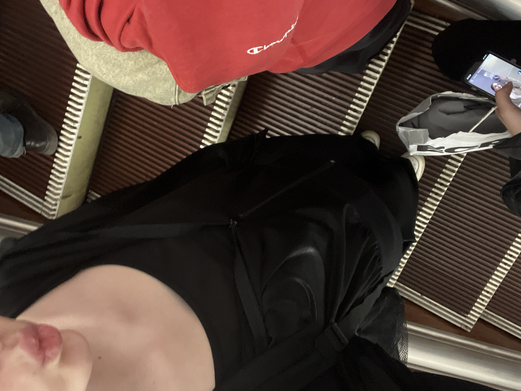 The image provides a high-angle, downward view of people on an escalator, likely within an indoor public space in Saint Petersburg, Russia.
In the foreground, the photographer's upper torso, including a mouth and chin, is visible, dressed in black clothing. Directly ahead, another individual is seen from above, wearing a red sweatshirt featuring the white "Champion" logo prominently on the front. This person's lower half is covered by light grey fabric, possibly trousers or a skirt, and a dark shoe is visible on the left side of the frame. To the right, a third person is holding a black smartphone, which displays a video call or content featuring human faces. This individual is wearing dark clothing and white sneakers, with a white shopping bag or tote bag partially visible near their feet.
The escalator steps are dark brown with distinctive white ribbed edges, forming a clear pattern. A metallic silver escalator handrail is visible along the right side of the frame. The lighting suggests an indoor setting with artificial illumination. The scene captures the everyday activity of people riding an escalator, with one person engaged with their mobile device.