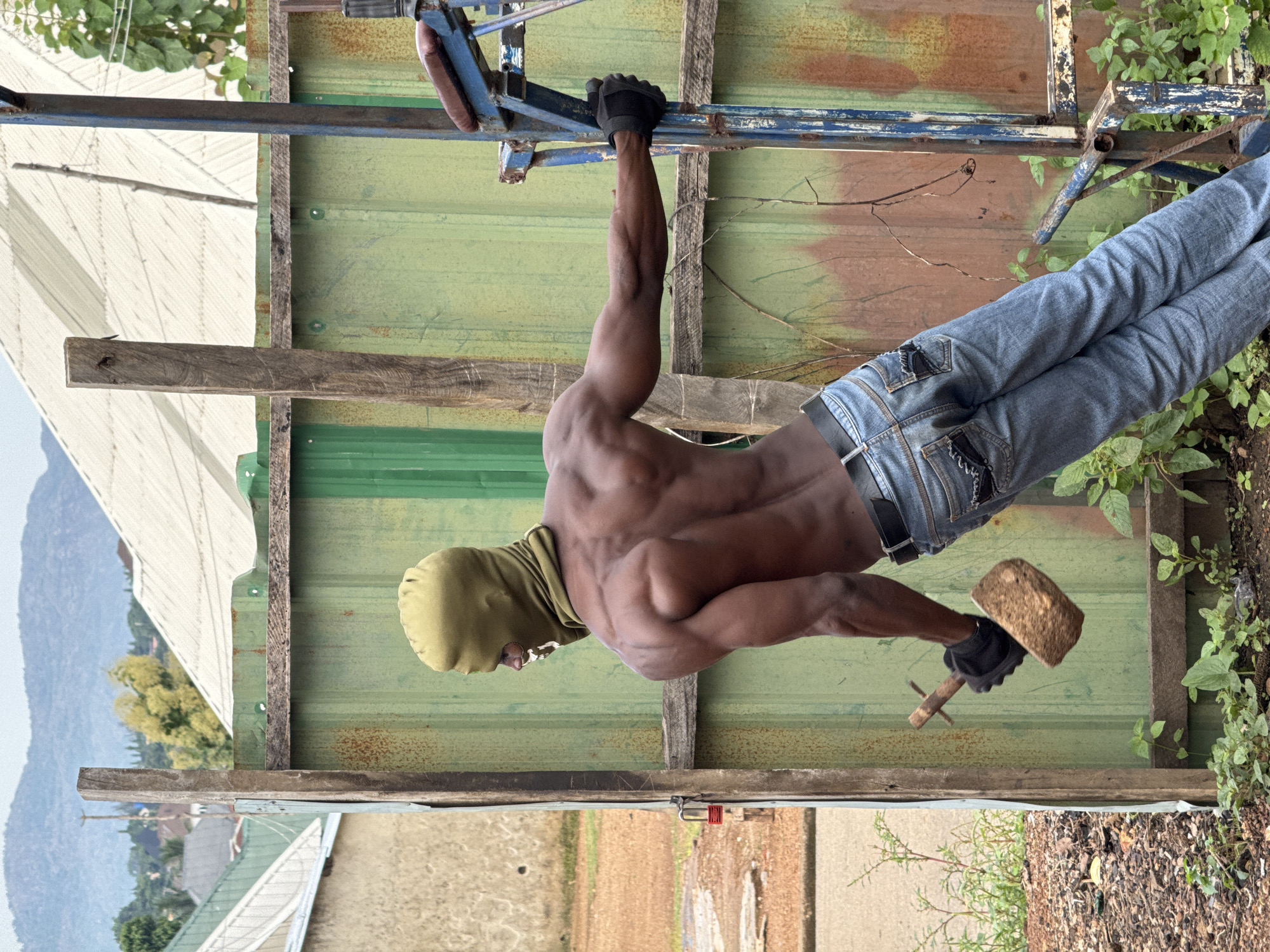 The image depicts a muscular Black man, shirtless and wearing a light olive green balaclava that covers his head and neck, with only his eyes and mouth visible. He is wearing blue jeans and black gloves. He is holding a mallet in his right hand and appears to be performing an exercise. The man is positioned outdoors in what appears to be an industrial or construction area. He is leaning against a tall, green corrugated metal wall, supported by a wooden post. To his right, a piece of blue exercise equipment is visible, with a padded seat. The ground is covered in dirt and debris, with some green vegetation growing around the base of the metal wall. In the background, there are structures with white, sloped roofs, and a hazy mountainous landscape under an overcast sky. The scene has a gritty, utilitarian aesthetic.