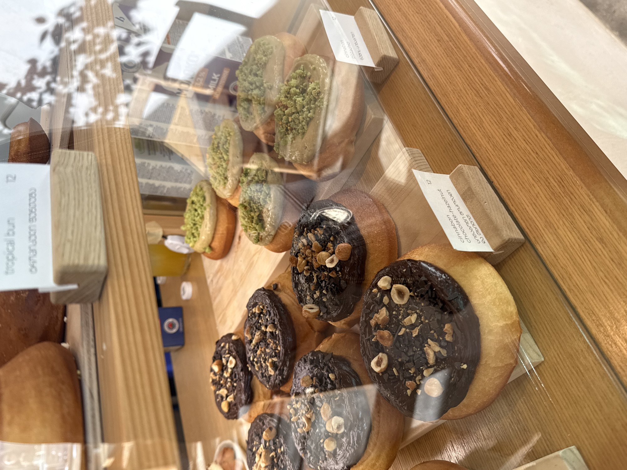 The image captures a close-up view of a display case filled with various baked goods, likely buns or donuts, in what appears to be a bakery or cafe in Batumi, Georgia. The pastries are arranged on wooden shelves inside a clear glass enclosure.
In the upper part of the display, several light-colored buns are visible, each topped with a generous amount of green-colored frosting or filling, likely pistachio, and sprinkled with crushed green nuts. Below these, a selection of darker buns is showcased, each coated in a rich, dark chocolate glaze and adorned with chopped hazelnuts.
Small white cards, held by wooden stands, provide descriptions for the pastries. One card identifies an item as "cinnabon pistachio" with Georgian script "ფსტა ფსტაშო" and the number "9". Another tag reads "cinnabon chocolate-hazelnut" accompanied by Georgian text "შოკოლად-თხილი" and the number "12". A third visible card lists "tropical bun" with Georgian script "ტროპიკული ფუნთუშა" and the number "12".
Reflections on the glass surface obscure some parts of the background but show blurry patterns, possibly foliage, and a partial view of a milk carton labeled "MILK БЕЗЛАКТОЗНОЕ" (Lactose-free milk) and "3,2%". The wooden elements of the display create a warm and inviting setting for the baked goods.