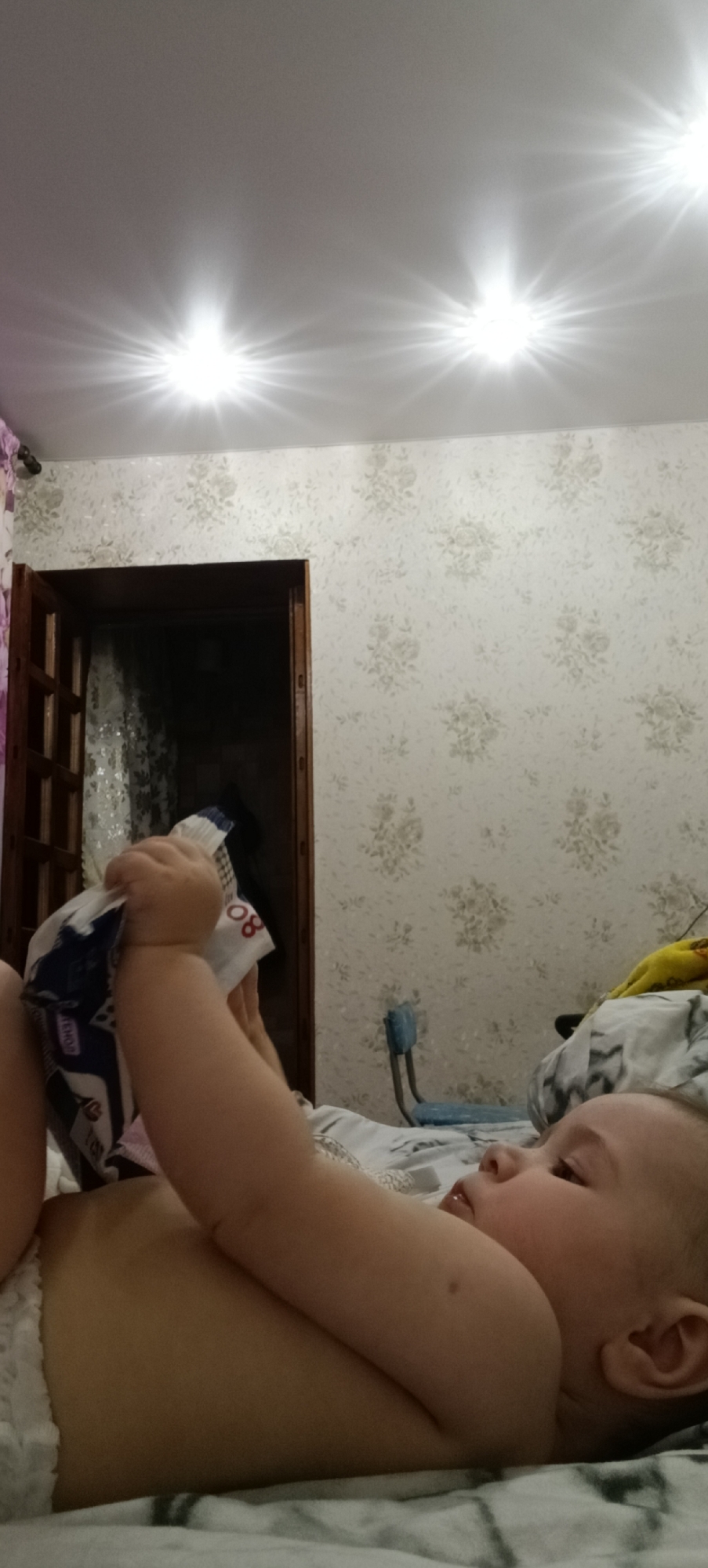 In an indoor scene, likely a bedroom, a baby is lying on a bed. The baby, seen from the side, is wearing a diaper. The baby's hands are raised, and they are holding a package. The room has wallpaper with a floral pattern and a ceiling with multiple bright lights. A partially open wooden door is visible on the left side of the image, leading to a darker space. A blue chair and some bedding are also present in the background. The specific location in the city of Хохлы, Russia, is not discernible from the visual information provided.