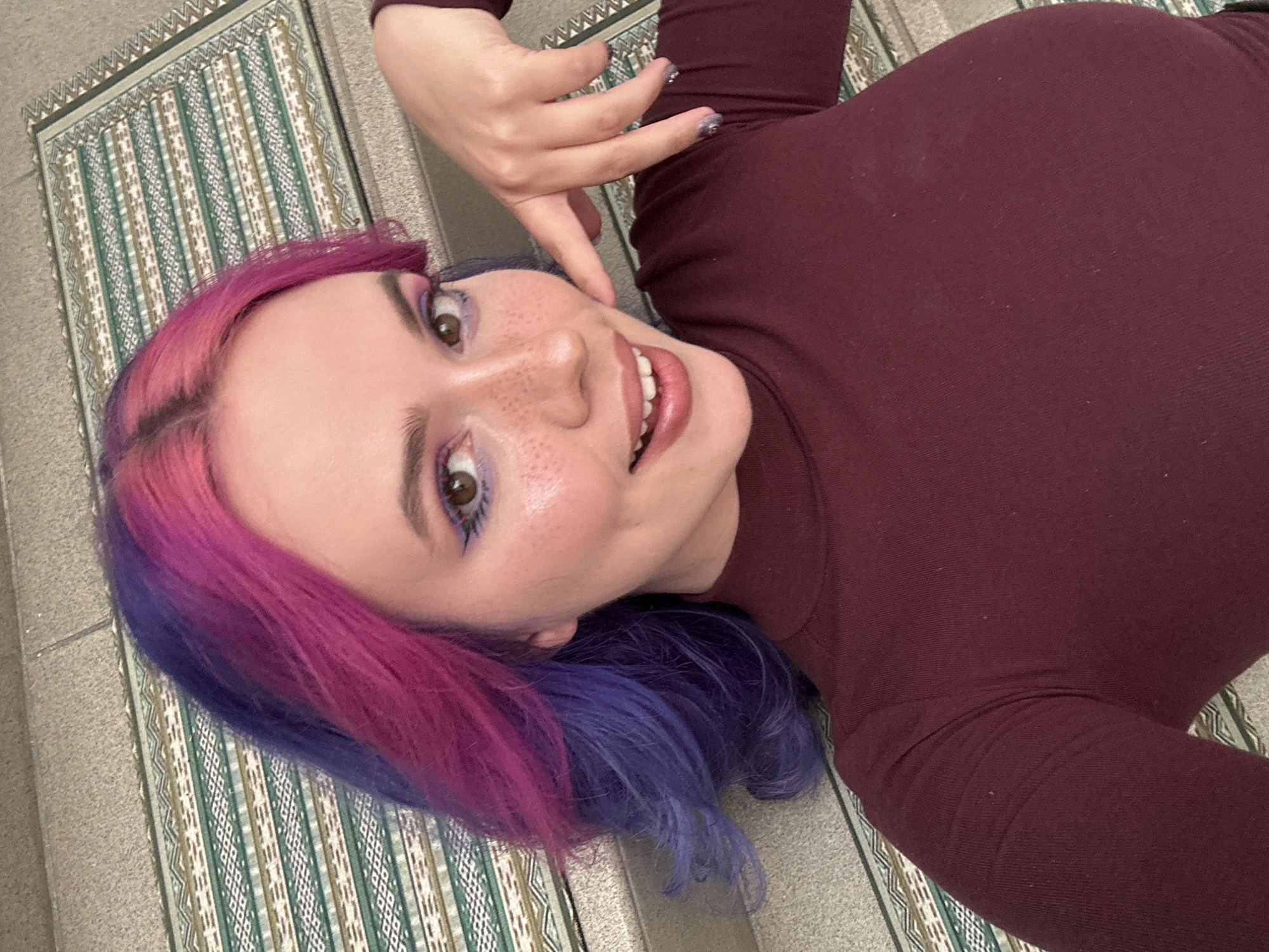 A vibrant portrait captures a young individual with distinct two-tone hair, featuring bright pink on the upper half flowing into purple at the ends. The person is lying down on their back, angled diagonally in the frame, and looking directly at the viewer with a wide, open-mouthed smile, revealing teeth. Their expression is cheerful and playful. They are wearing a form-fitting, long-sleeved top in a deep maroon color. Notable facial details include prominent purple eyeshadow and drawn-on freckles across the nose and cheeks. One hand is gently placed near their chin, showcasing glittery nail polish on manicured nails. The individual is positioned on a patterned mat or rug, characterized by alternating stripes of green, white, and brown geometric designs. A plain, light gray floor surface is visible next to the mat. The setting appears to be indoors within the City of Zagreb, Croatia, likely a casual moment or selfie. No visible text is present in the image.