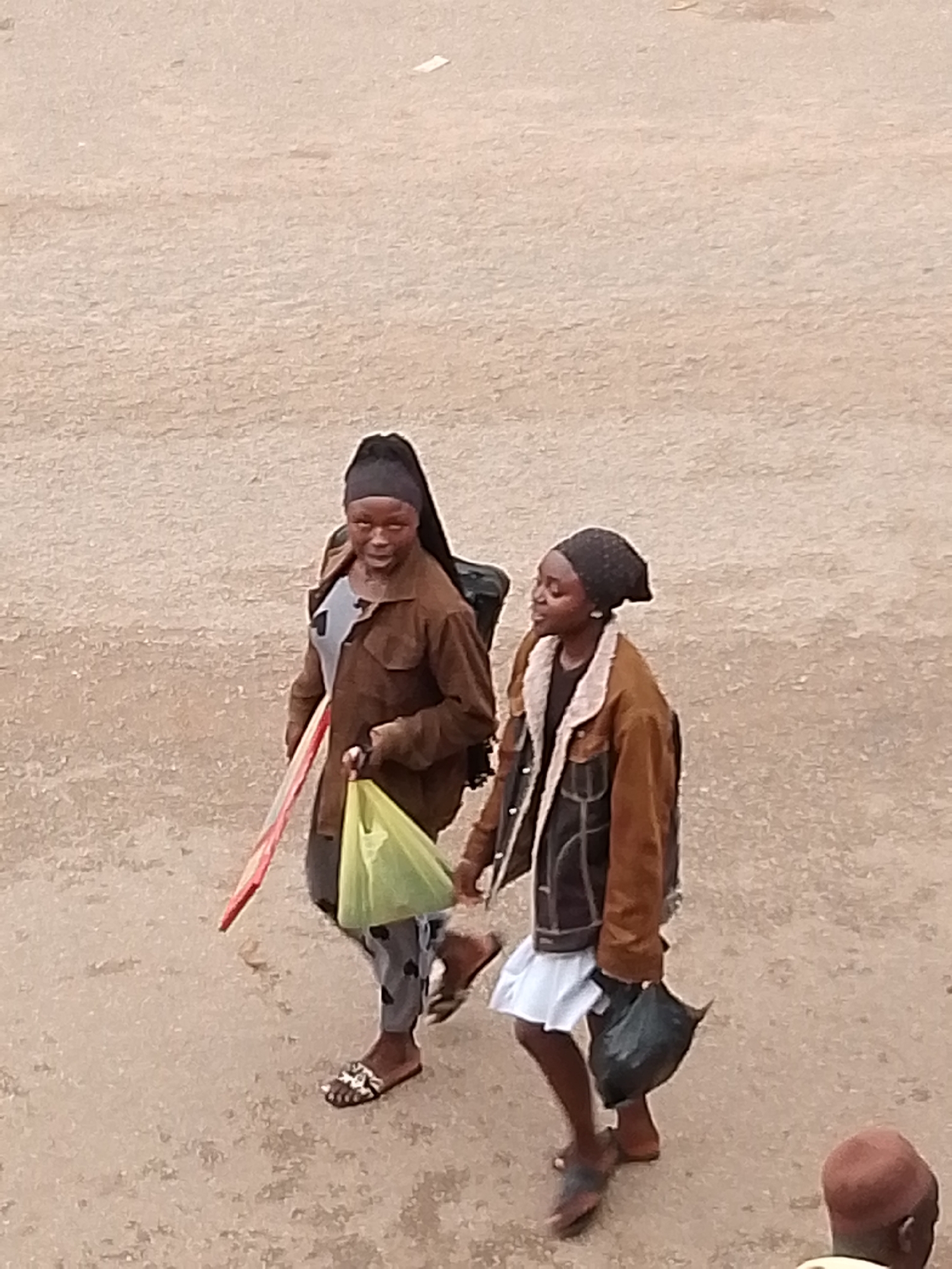 Two young women are walking on a dusty, light brown, unpaved surface in Bukuru, Nigeria. The woman on the left is wearing a brown jacket over a grey top and patterned grey pants, with a black headscarf and dark sandals. She is carrying a bright yellow plastic bag in her left hand and a flat, red and white object in her right hand. The woman on the right is wearing a brown jacket with a shearling collar over a dark top, a white skirt, and dark sandals. She is carrying a black plastic bag in her right hand and has a black headscarf on. Their faces are visible, and they appear to be looking forward. The background is a uniform, textured dusty ground. The image is taken from a high angle, looking down on the subjects.